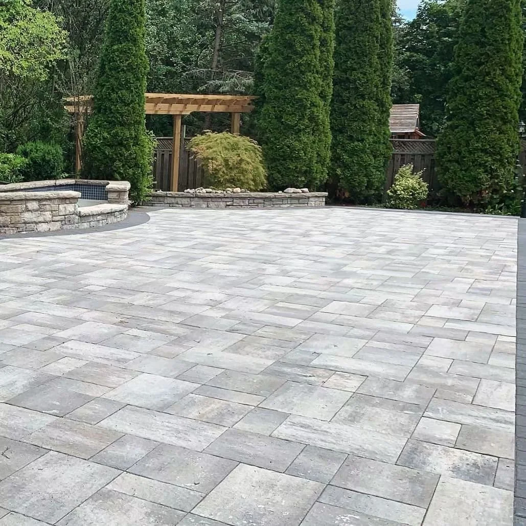 A spacious backyard patio with grey stone tiles, a stone hot tub on the left, and a landscaped garden with tall green trees and plants in the background.