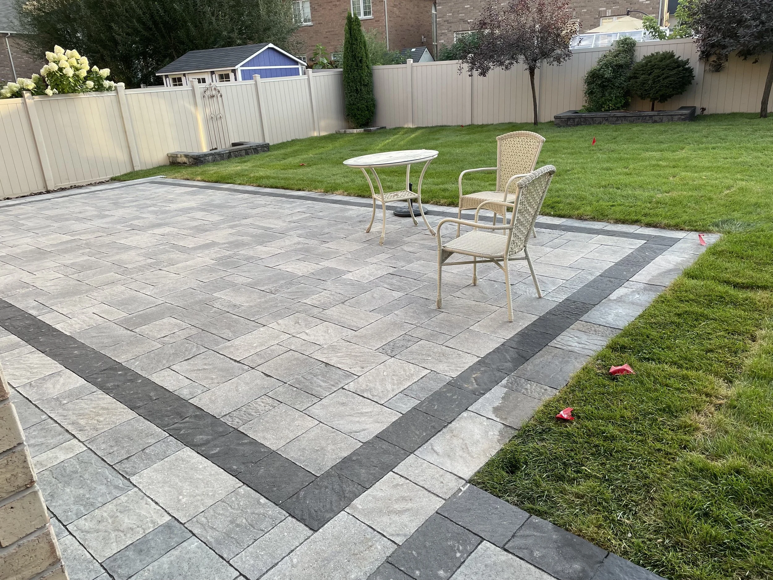 Backyard patio with gray stone tiles and a border of darker stones, furnished with two wicker chairs and a small round table, surrounded by green grass, garden beds, trees, a white fence, and neighboring houses in the background.