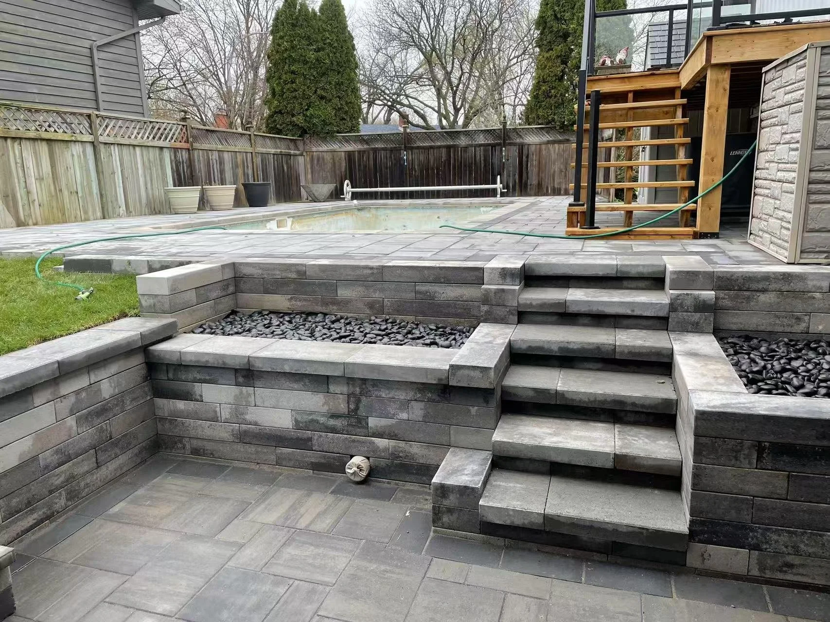 Backyard with a raised patio area, steps, and a swimming pool, fenced with a wooden fence, with trees in the background and potted plants along the fence.
