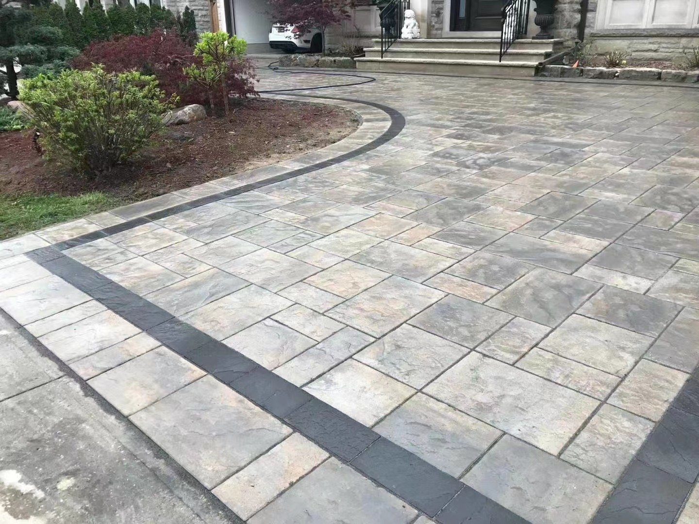 A paved driveway with a border of darker stones, leading to a front porch with steps and columns, and a garage in the background. There are plants and shrubs on the side.