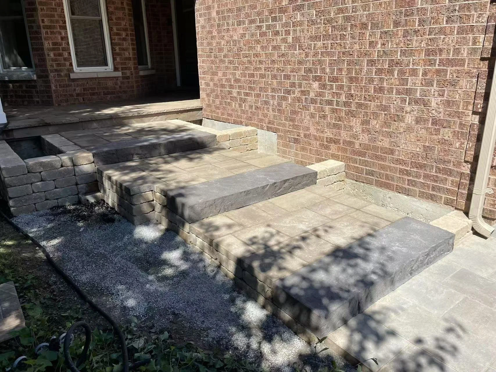 Newly constructed outdoor brick and stone patio with stairs leading to house entrance, partially shaded by tree shadows.