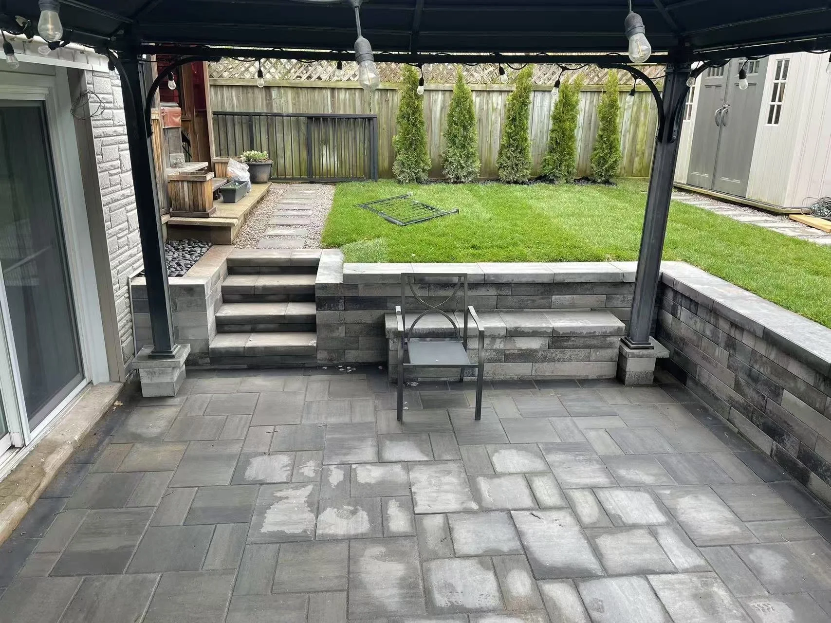 Backyard patio with gray tile flooring, steps leading up to a grassy area, and a black metal chair on the patio under a gazebo. The yard has green grass, a row of tall trees, and a wooden fence.
