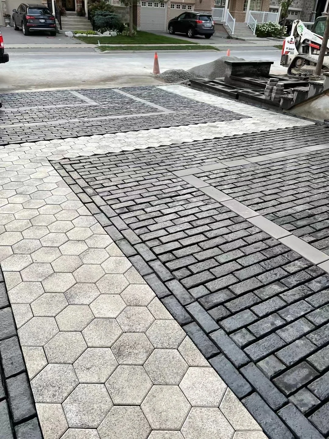 A sidewalk with different paving patterns, including white hexagonal tiles, dark brick, and concrete strips, leading to a construction site with a compact machine, dump pile, and orange traffic cones. Residential houses and parked cars are visible in