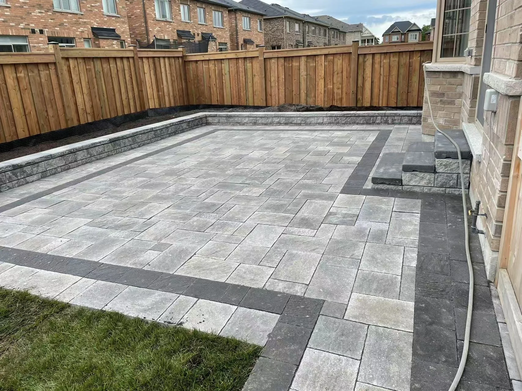 A finished backyard patio with grey concrete pavers, a border of dark grey pavers, a small grassy area on the left, a wooden fence in the background, and steps with black pavers leading up to a house with brick exterior.