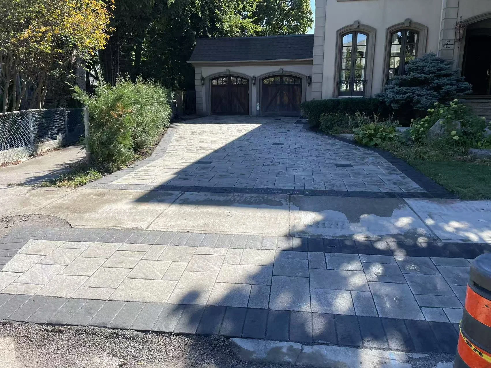 Newly paved driveway with patterned concrete in front of a house, with trees and bushes on sides, and a gated garage in the background.
