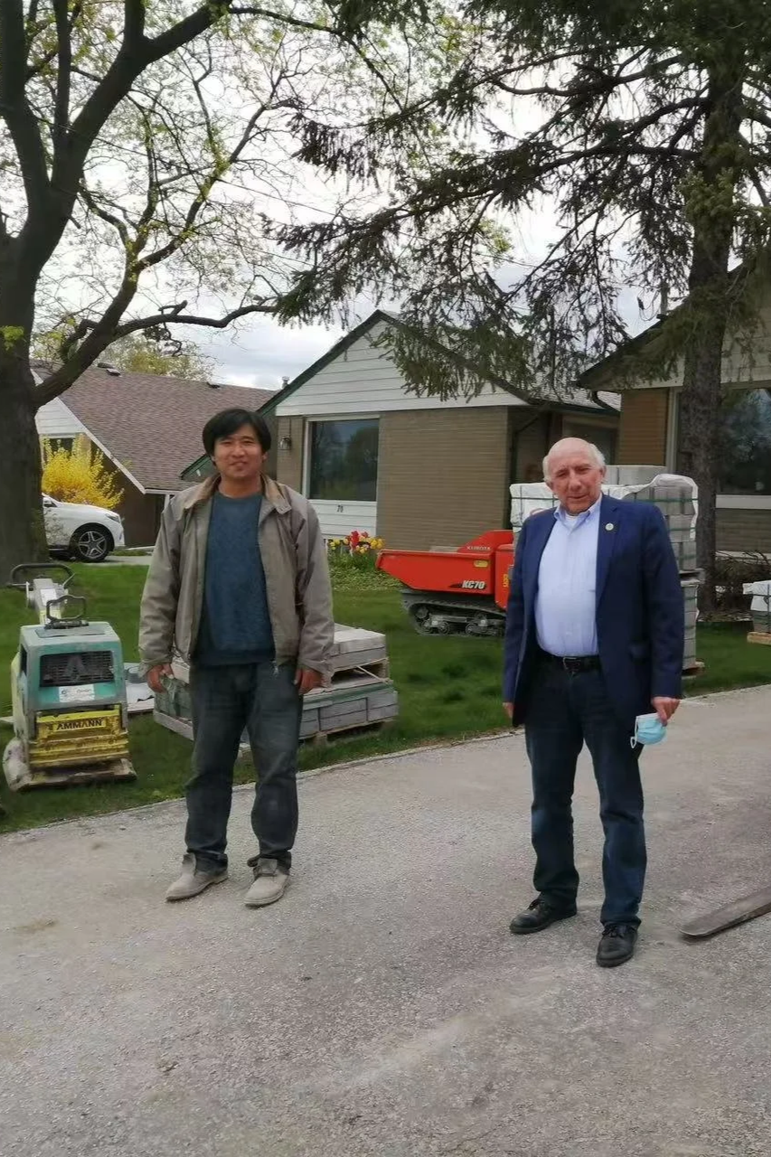 Two men standing outside in front of a house, one was in casual attire and the other in a suit, with construction equipment and a tree in the background.