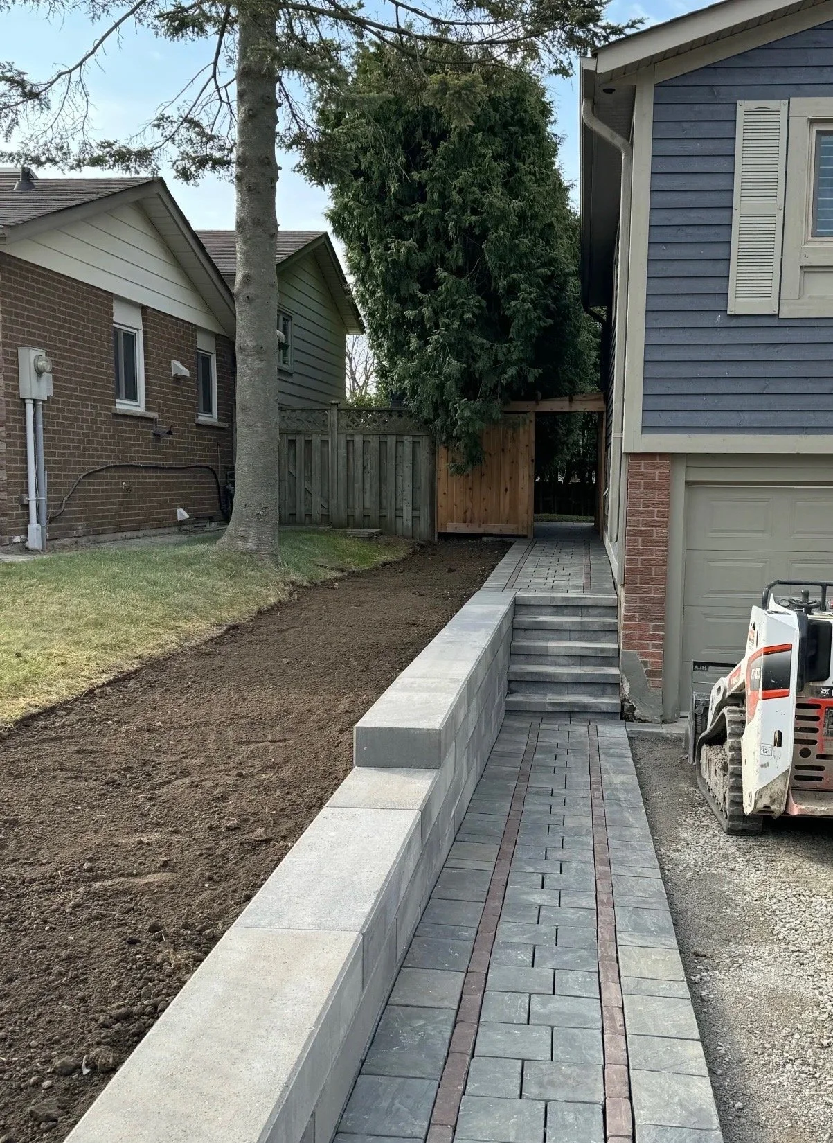 Sidewalk and stairs leading to a backyard, with newly laid brick and concrete edging, adjacent to a house with blue siding, a garage, and a construction machine.