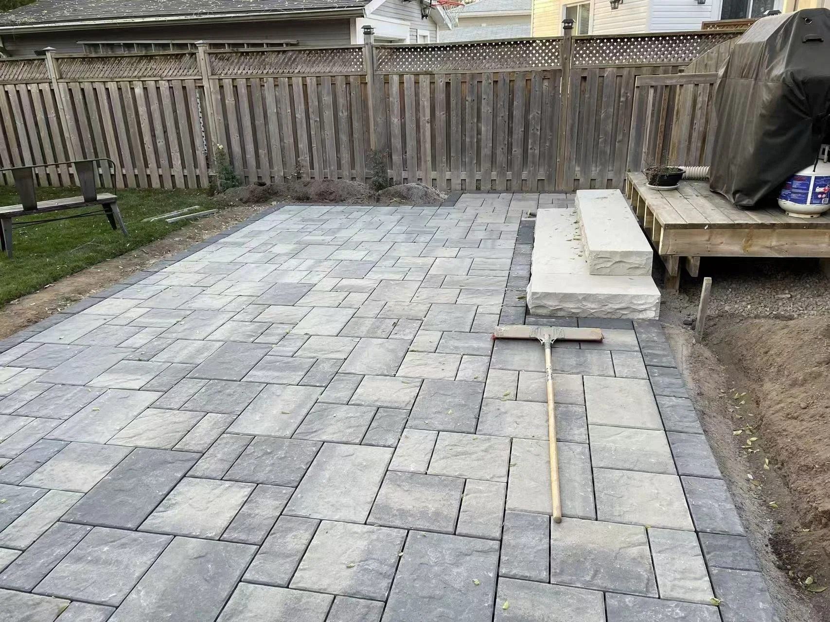Newly paved concrete patio with stone tiles, surrounded by a wooden fence, a bench, and construction tools, including a broom, with some construction materials on a wooden deck.