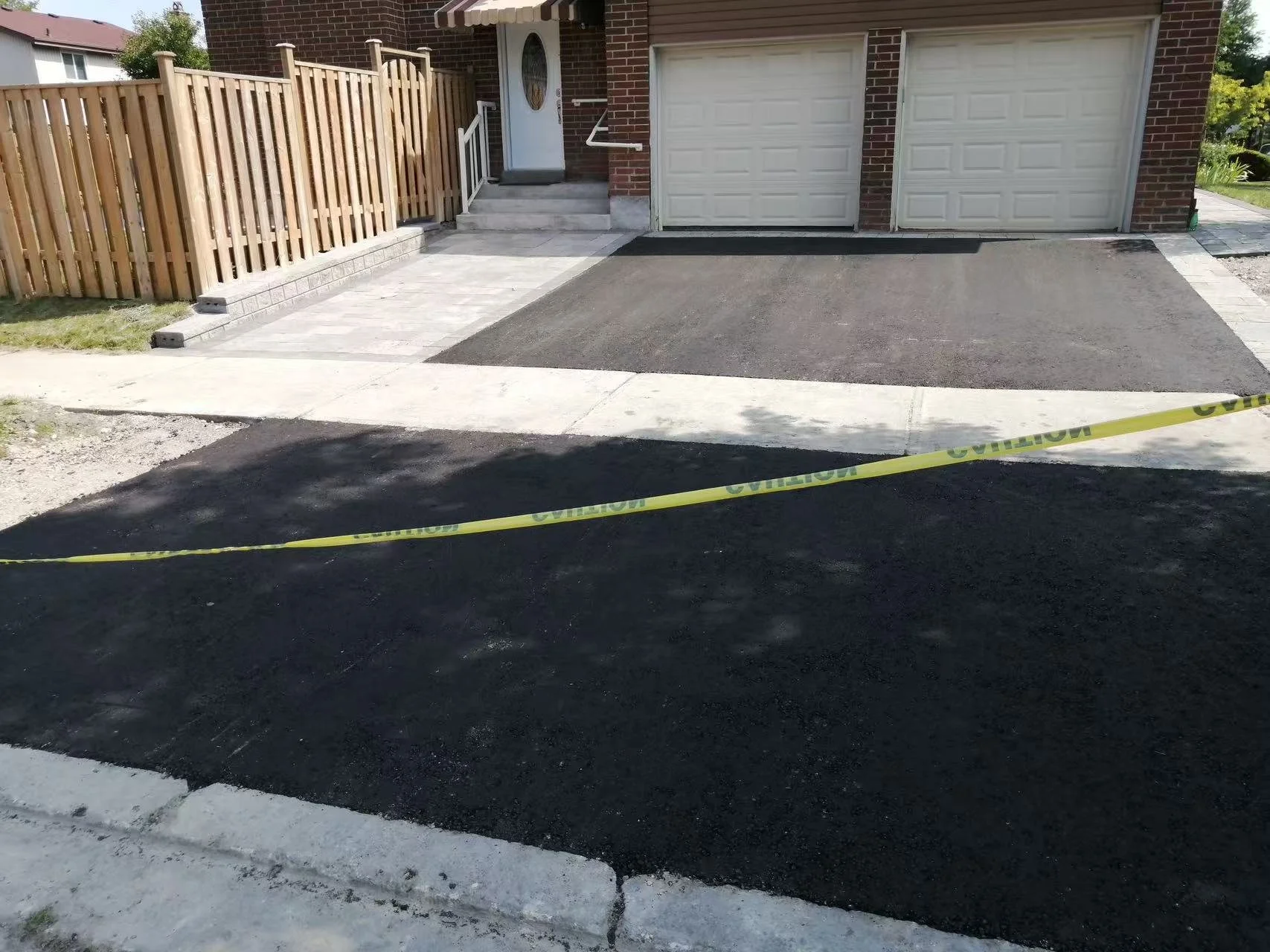 Recently paved driveway and patio areas in front of a brick house with a white garage door, a front door, a wooden fence, and caution tape across the freshly laid black asphalt.