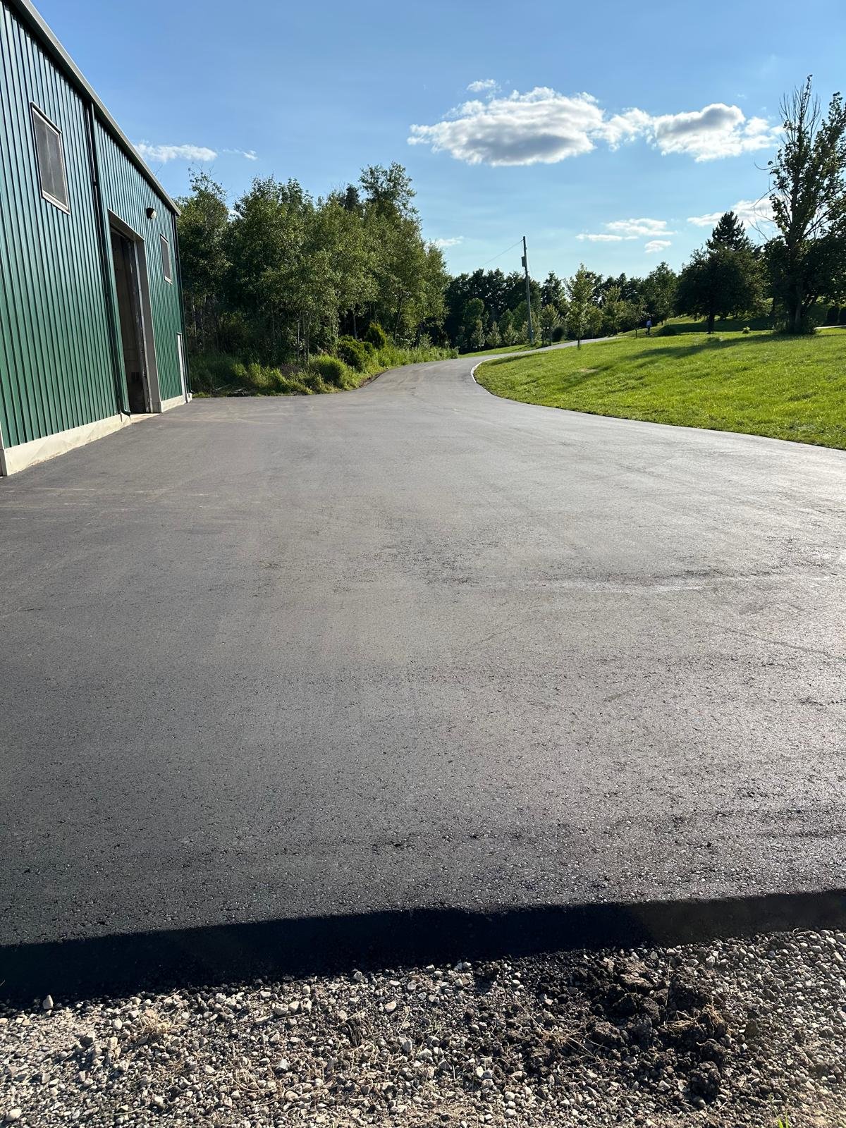 A paved driveway curves around a green grassy area with trees under a partly cloudy blue sky.