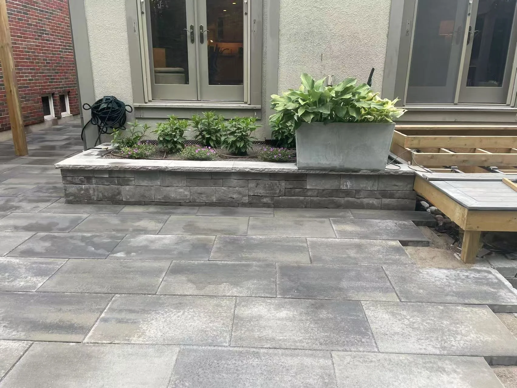 A backyard patio with grey stone tiles, a raised flower bed with dark brick border, and large planters with green plants in front of a house with beige textured walls and glass-paneled doors.