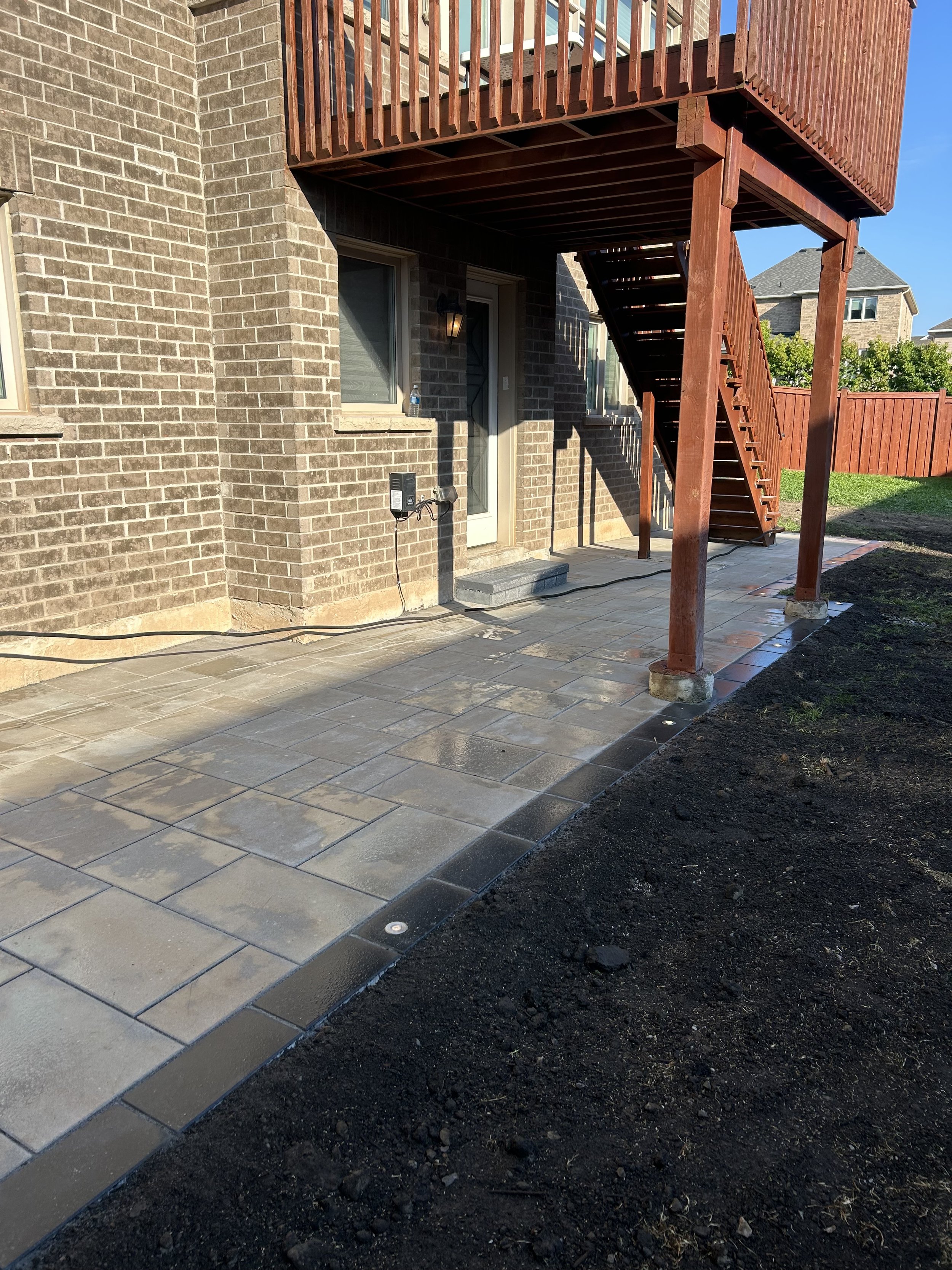 Backyard patio with newly installed stone pavers, brick house wall with door and window, and a wooden staircase leading to an upper deck, surrounded by a red wooden fence.