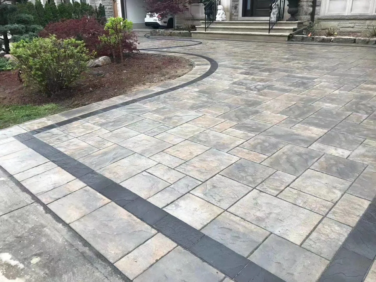 A paved driveway with a border of darker stone, curving toward a house entrance with steps, a white statue, and a gateway. The driveway is adjacent to a landscaped area with bushes and small trees.