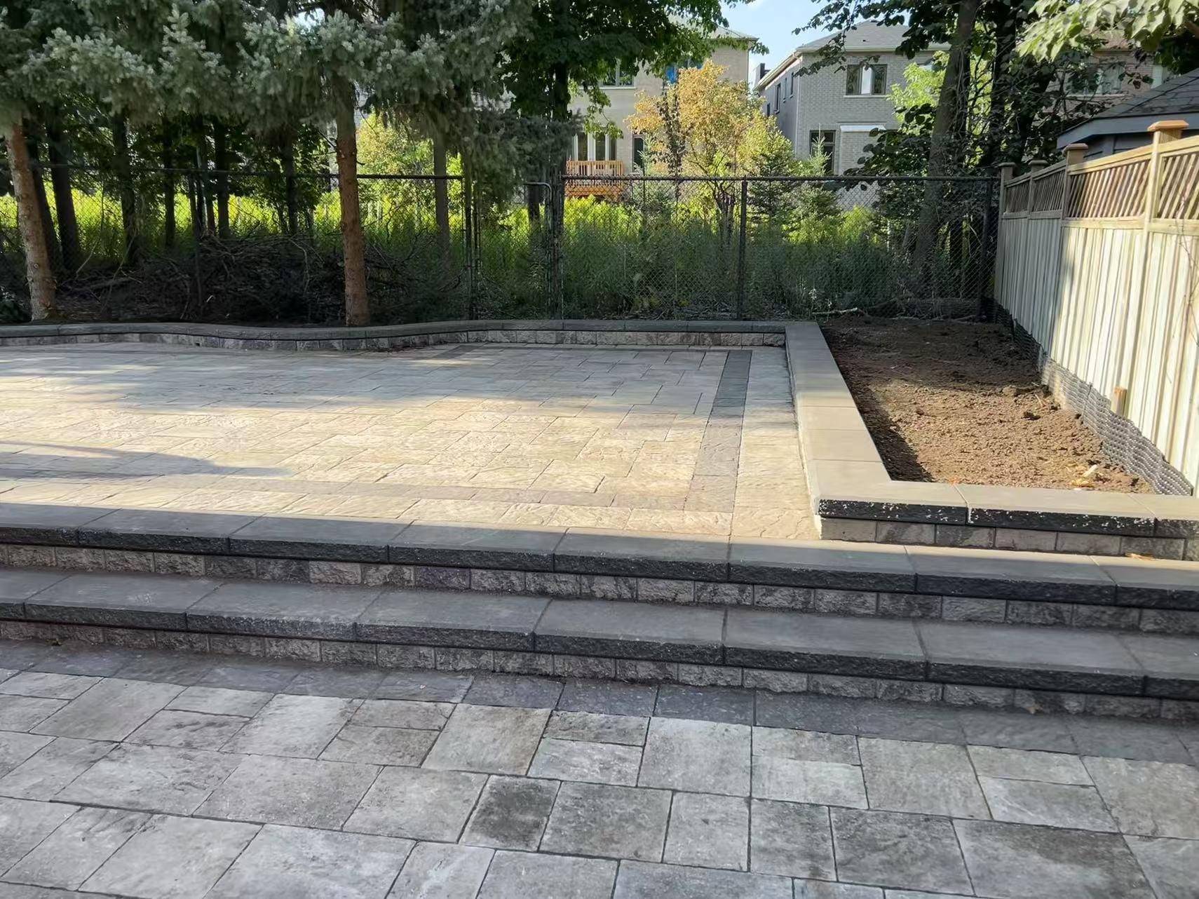 Backyard with freshly paved stone patio, stairs leading up to it, and a garden bed with fencing, surrounded by trees and residential houses.