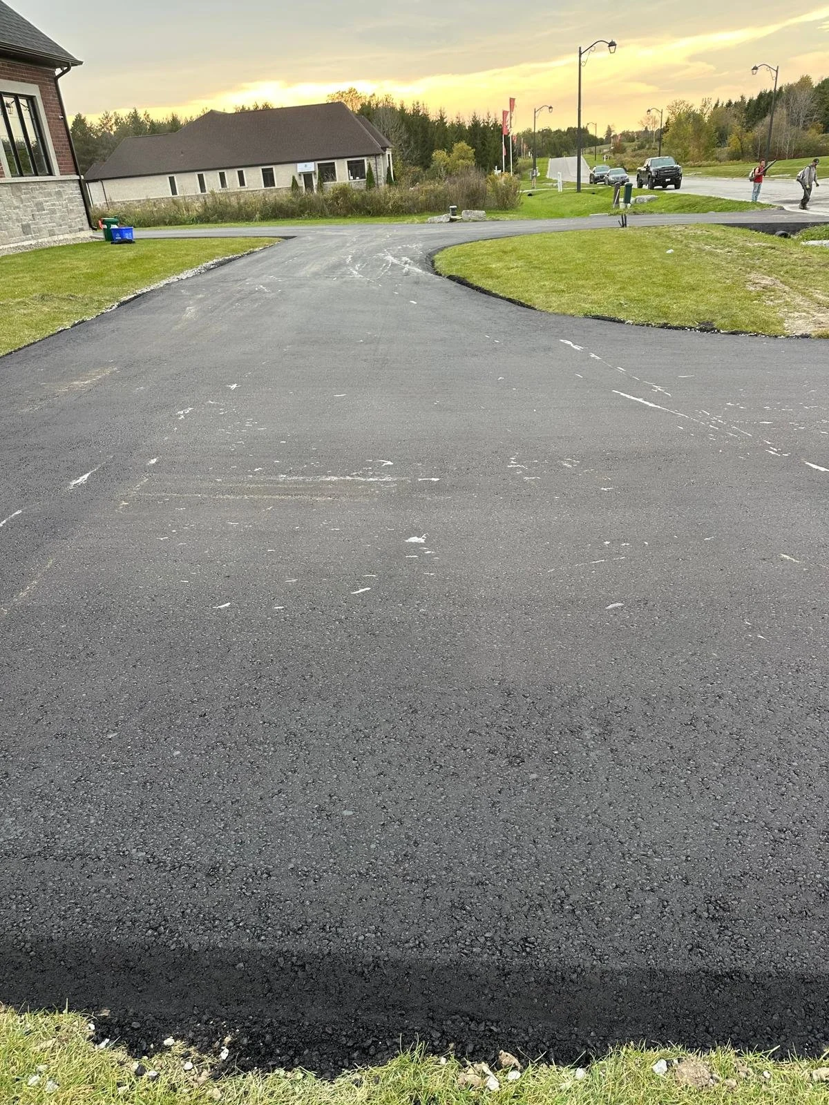 Freshly paved black asphalt driveway with some white markings, bordered by grass on both sides, leading to a residential area with houses and a street with cars in the distance at sunset.