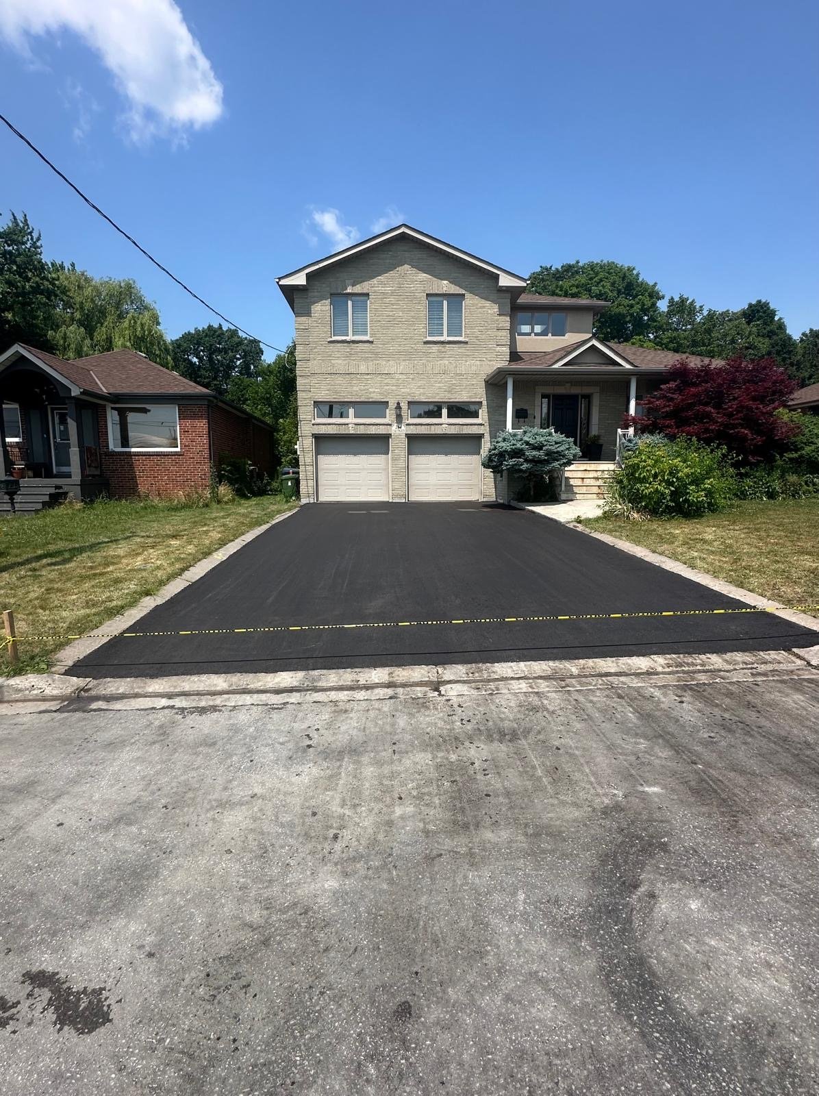Newly paved black asphalt driveway leading to a house with two garage doors, adjacent to neighboring homes, under a blue sky with a few clouds.