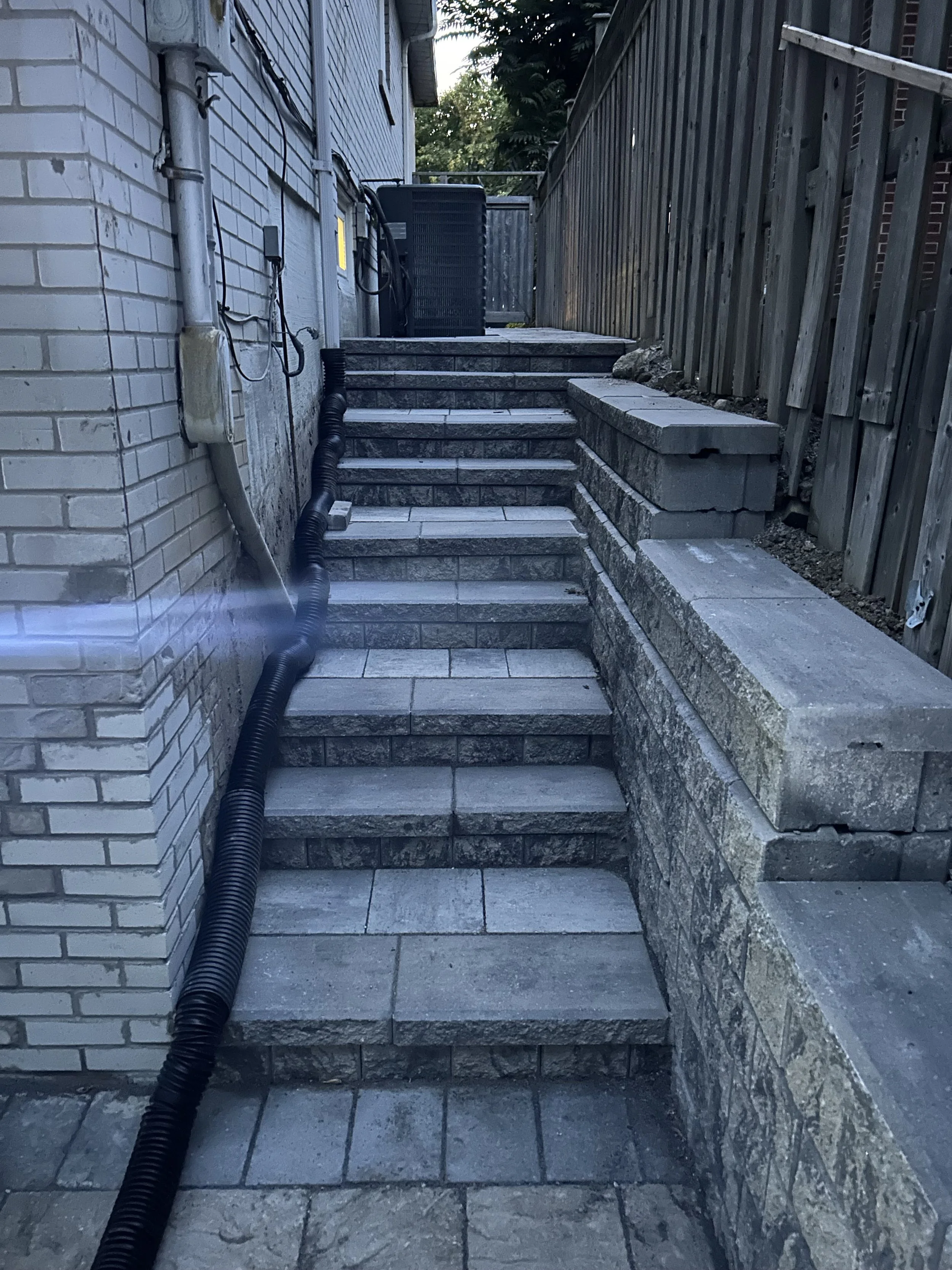 StoneSteps leading up to a backyard, with a brick wall on the left, a wooden fence on the right, and an HVAC unit at the top. A black drainage pipe runs along the side of the house.