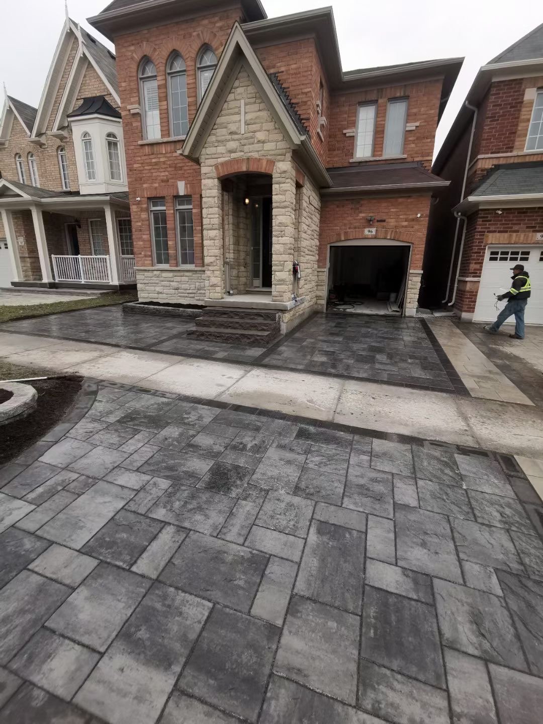 Front view of a newly paved black and grey brick driveway in front of a two-story brick house under construction, with a worker standing nearby.