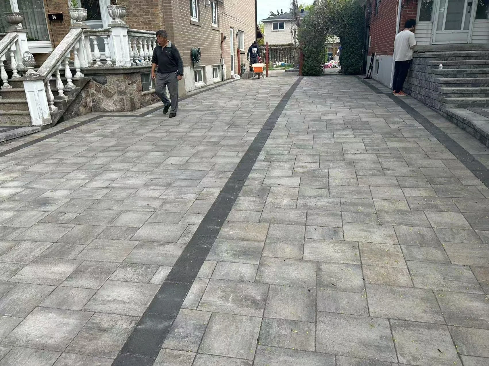 Residential backyard with a paved stone patio, black border stones, and stone stairs leading to a porch. Two people are standing on the patio, one near the house and one further back.