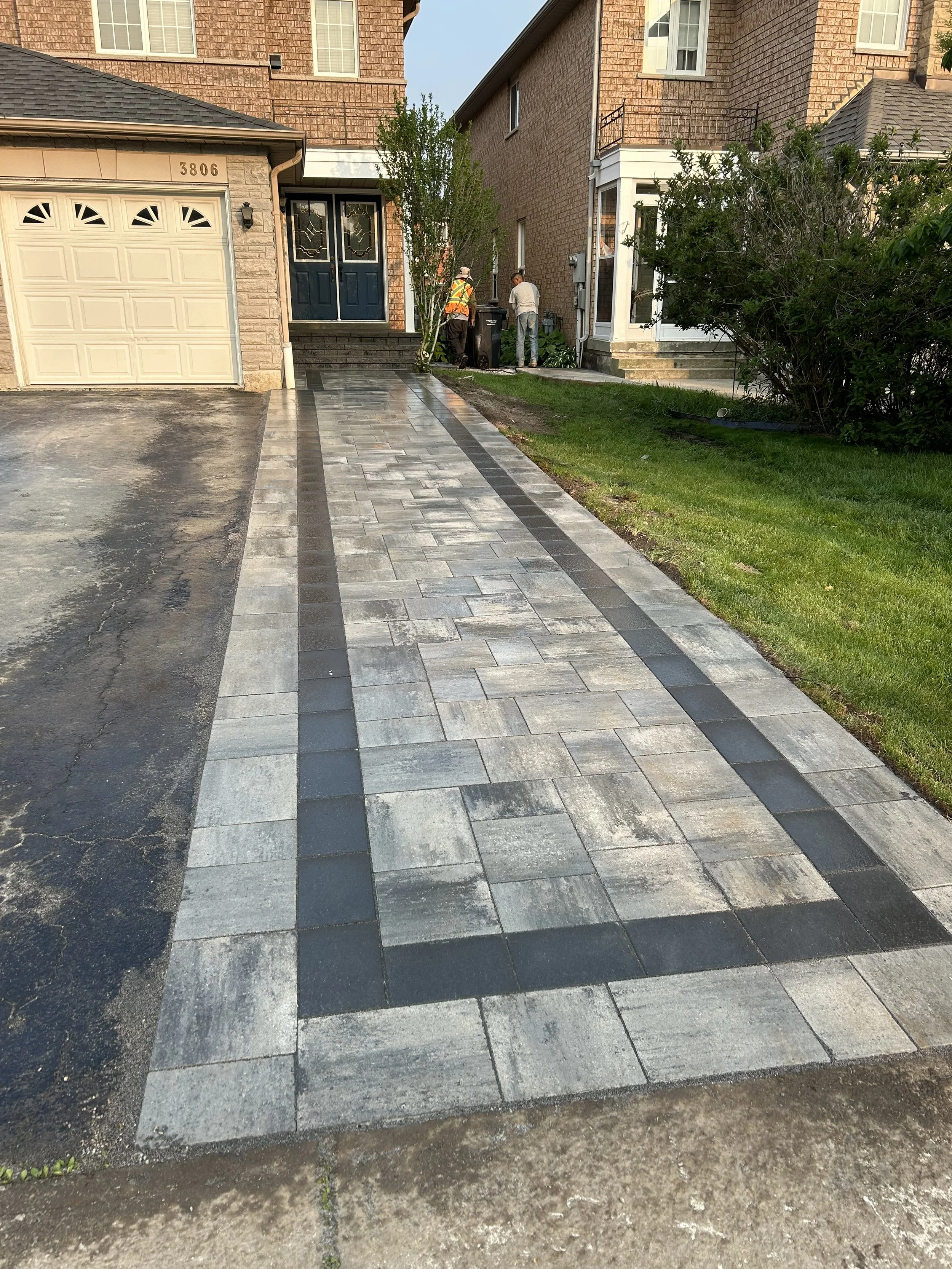Newly paved concrete driveway and walkway leading to the front entrance of a brick house. Two workers are near the door, engaged in work.