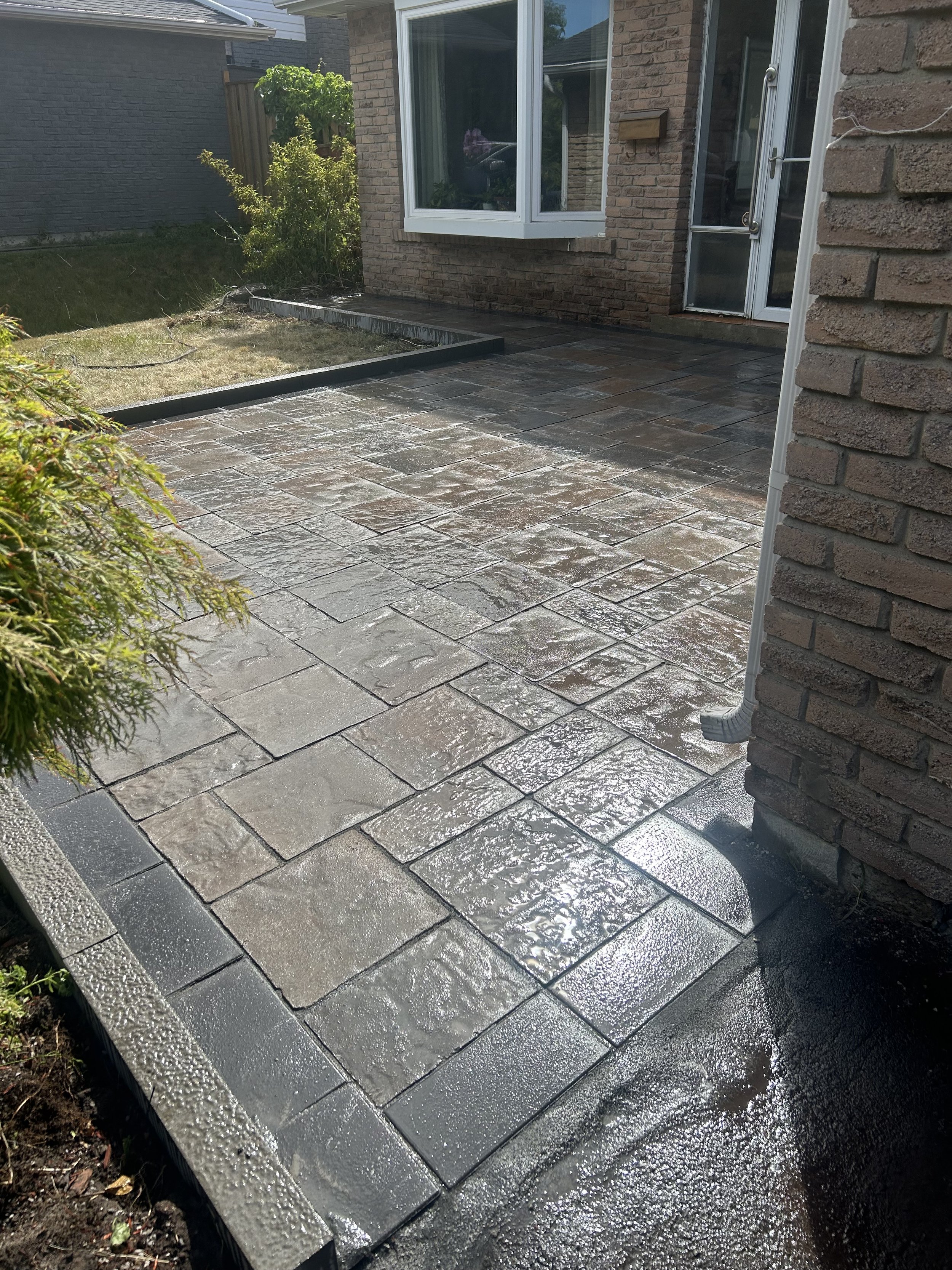 Clean, wet paver patio outside a brick house with a bay window and a glass door, surrounded by a garden with bushes and grass.