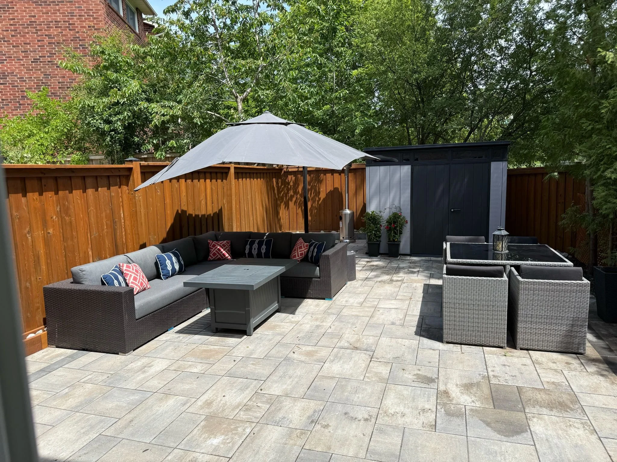 An outdoor patio area with a gray sectional sofa under a large umbrella, a gray coffee table, a black storage shed, and a dining table with six chairs, all surrounded by a wooden fence and green trees.