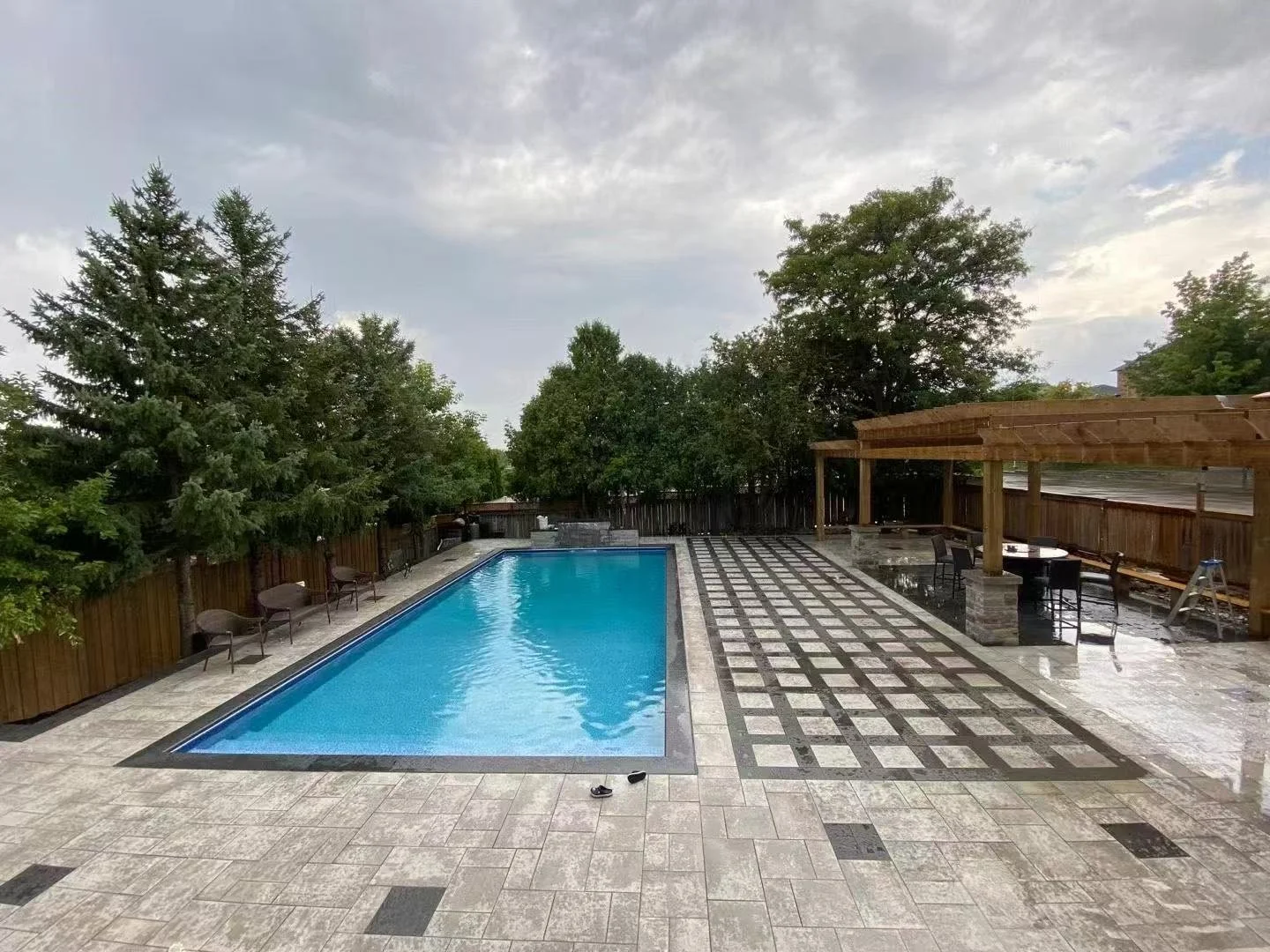 Backyard swimming pool with fence, patio, and seating area, surrounded by trees, under a cloudy sky.