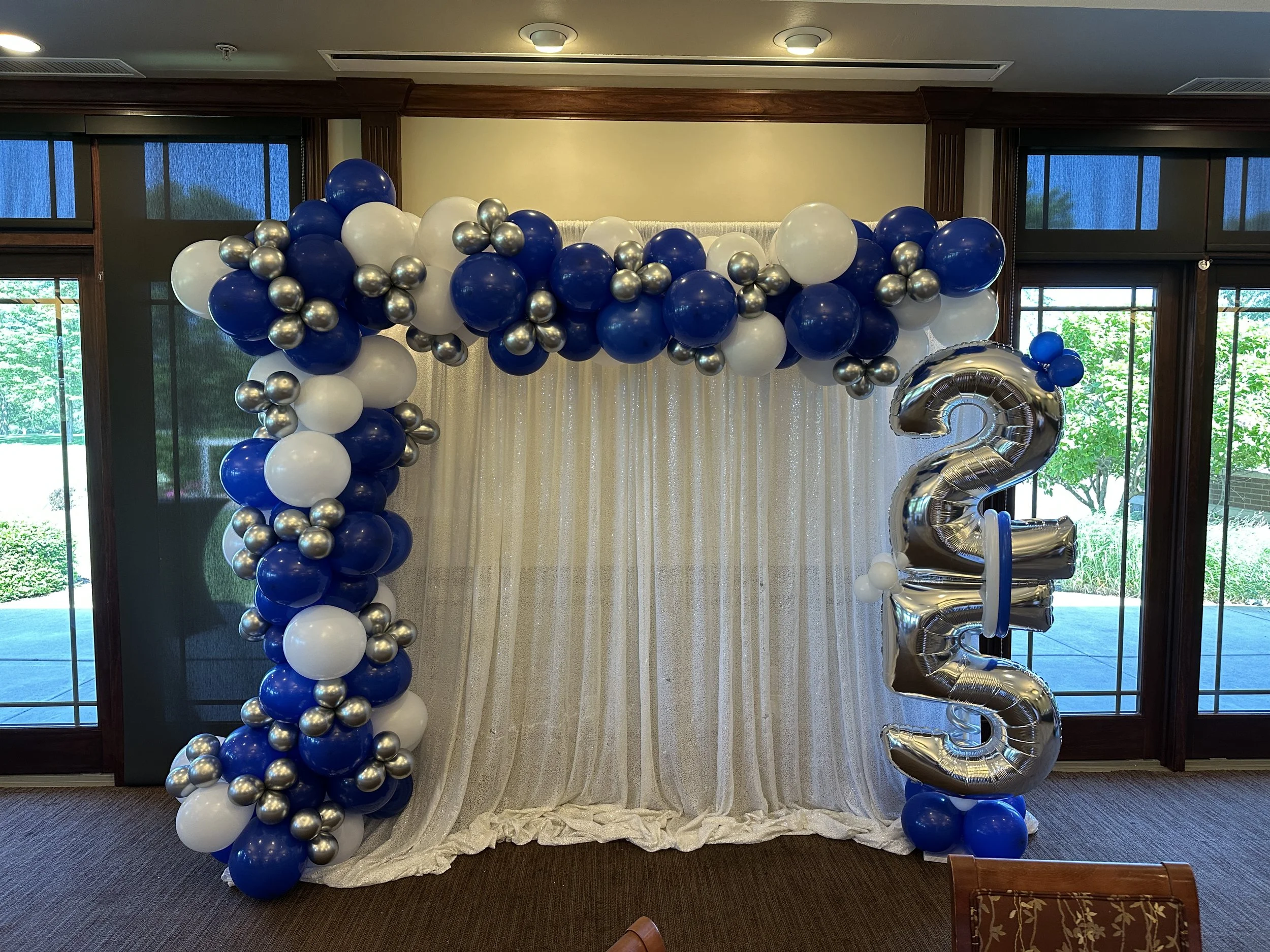 Balloon decoration with blue, white, and silver balloons forming an arch and the number 23 in silver foil balloons, set against a draped white backdrop inside a room with large windows.