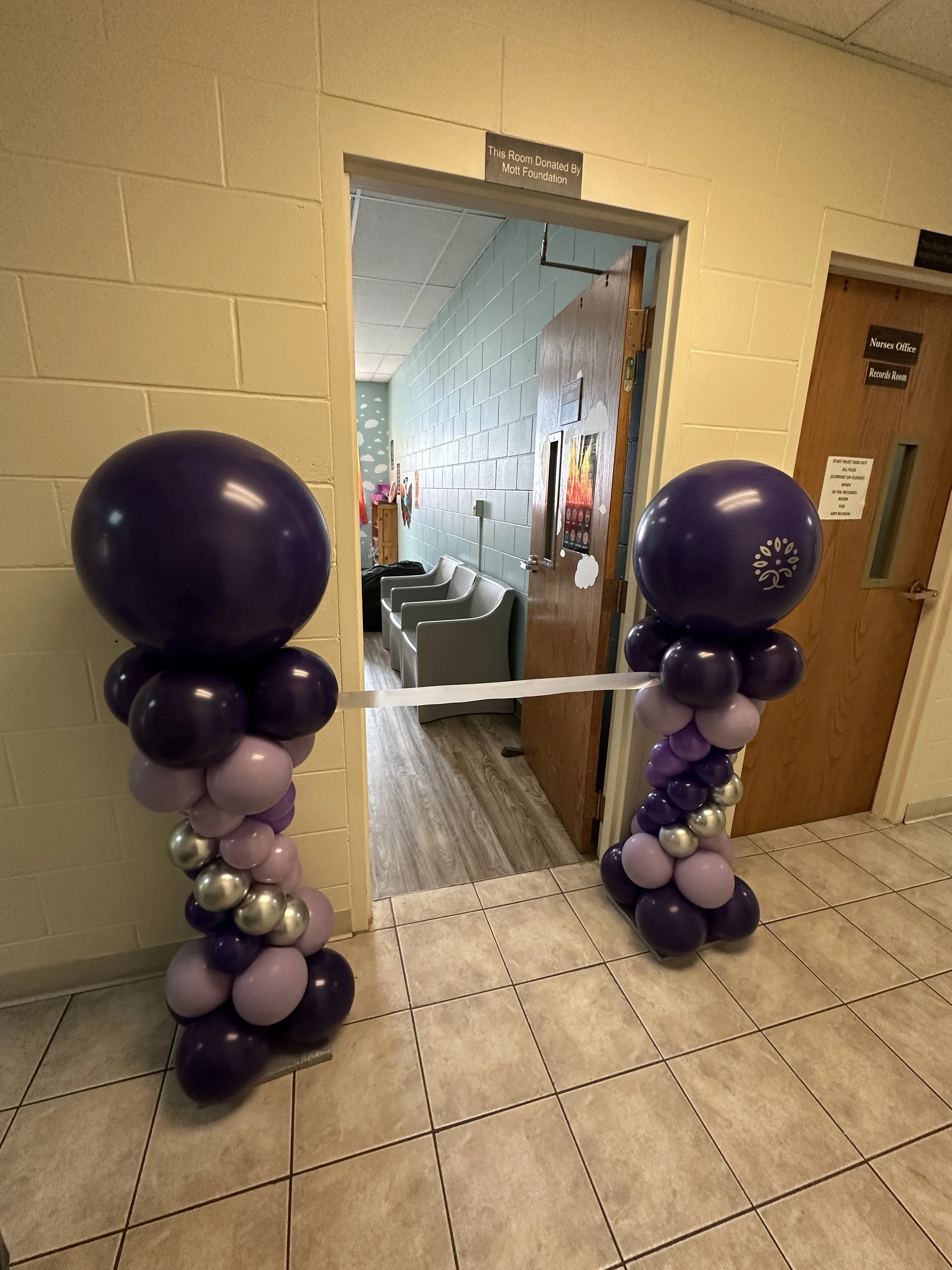 Balloon arch with purple, silver, and lavender balloonsdecorates an indoor doorway leading to a room, with a sign above the door reading 'This Room Donated By Mott Foundation'.