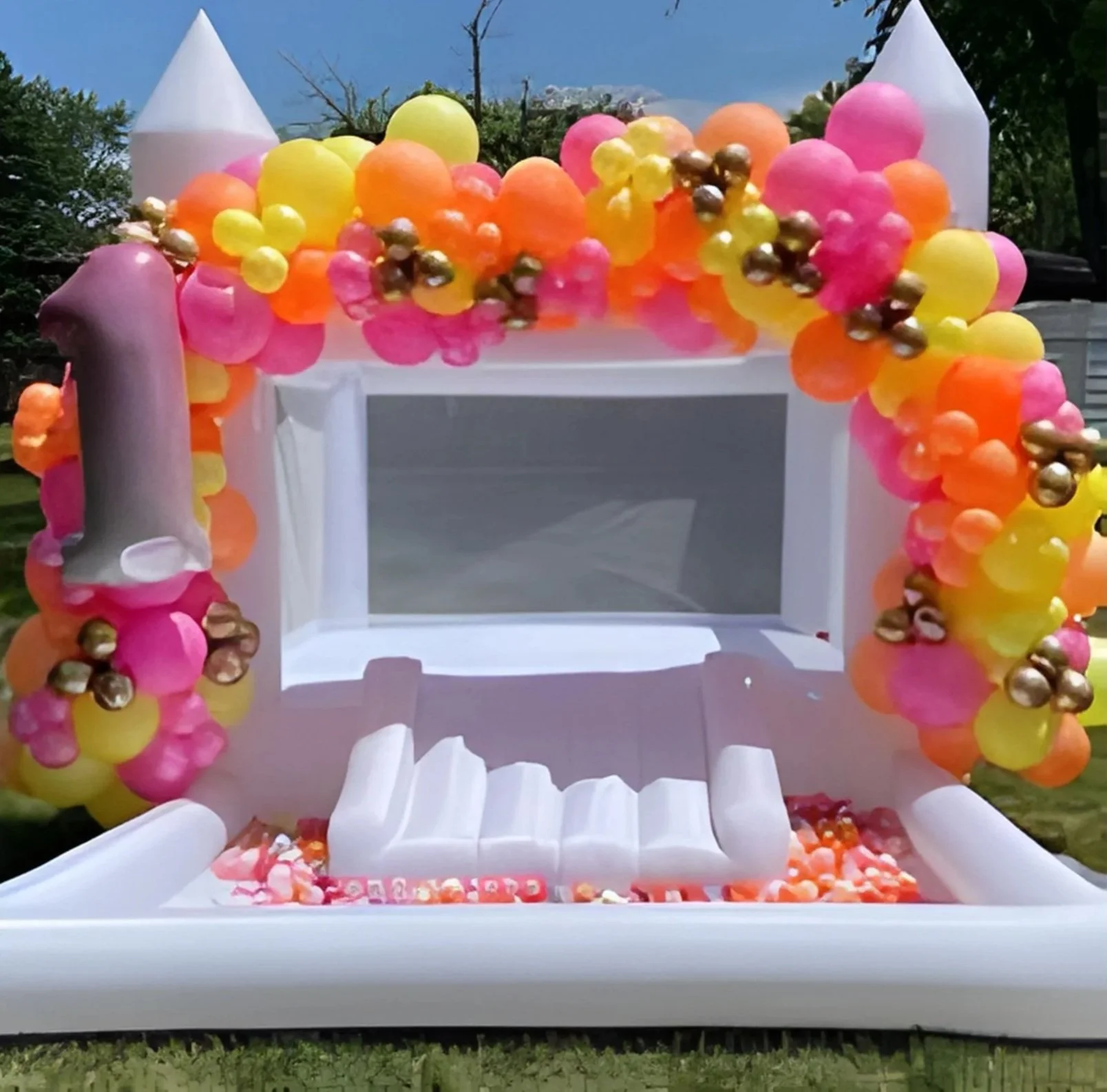 Inflatable outdoor birthday party decoration with a white castle and a balloon arch in pink, yellow, orange, and gold, with a large number 1 candle on the left.