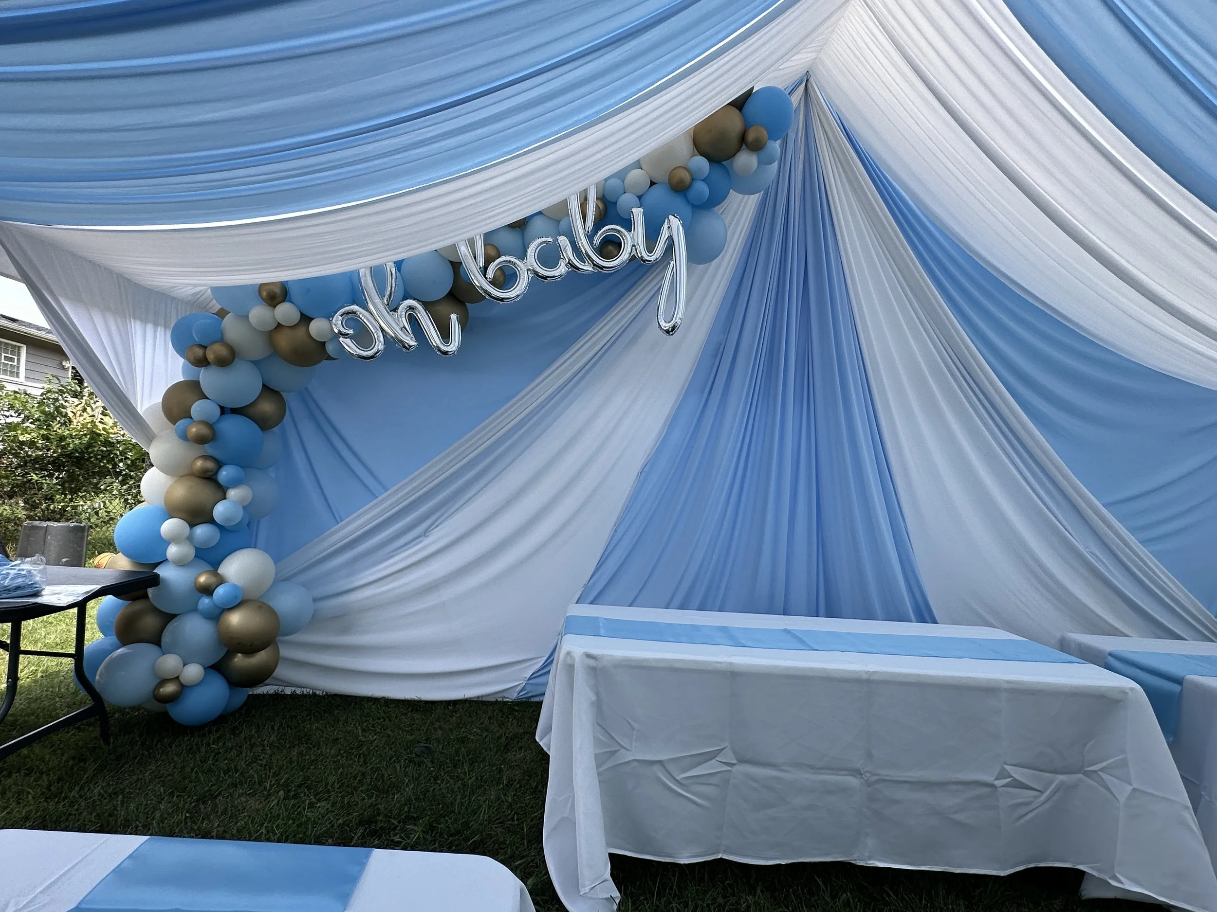 Baby shower setup with blue, white, and gold balloons, draped fabric, and a sign that says 'oh baby' in silver balloons.