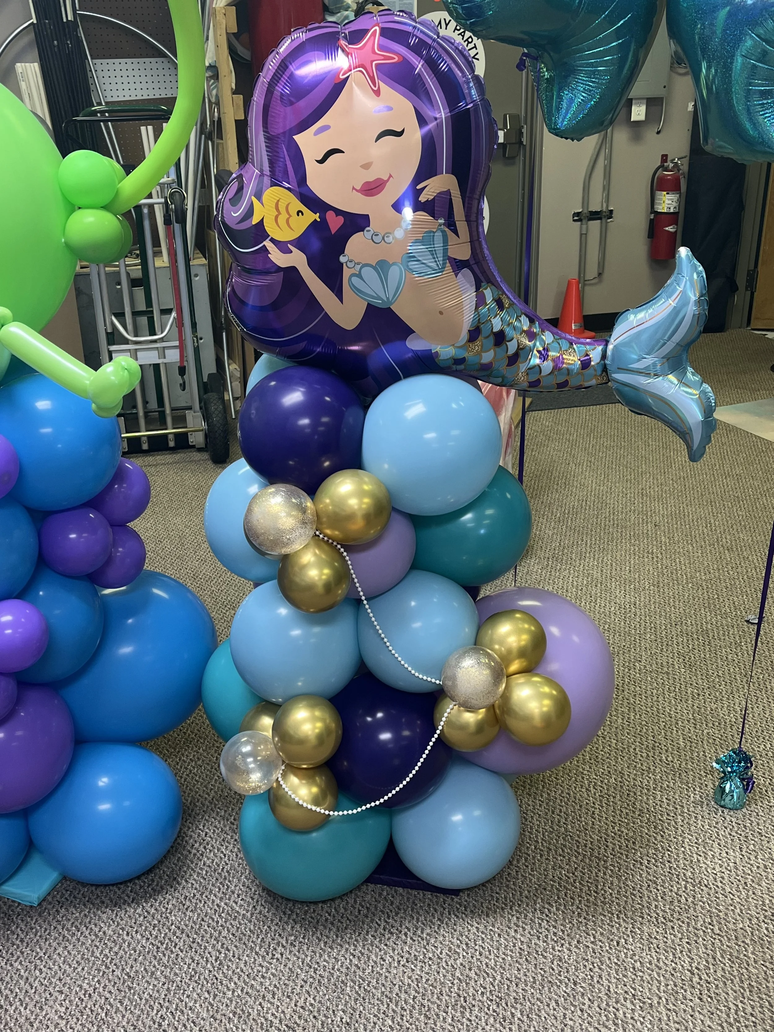 Balloon arrangement featuring a large mermaid-shaped balloon with purple hair, a yellow fish, and shells, surrounded by clusters of blue, purple, gold, and silver balloons on a carpeted floor.