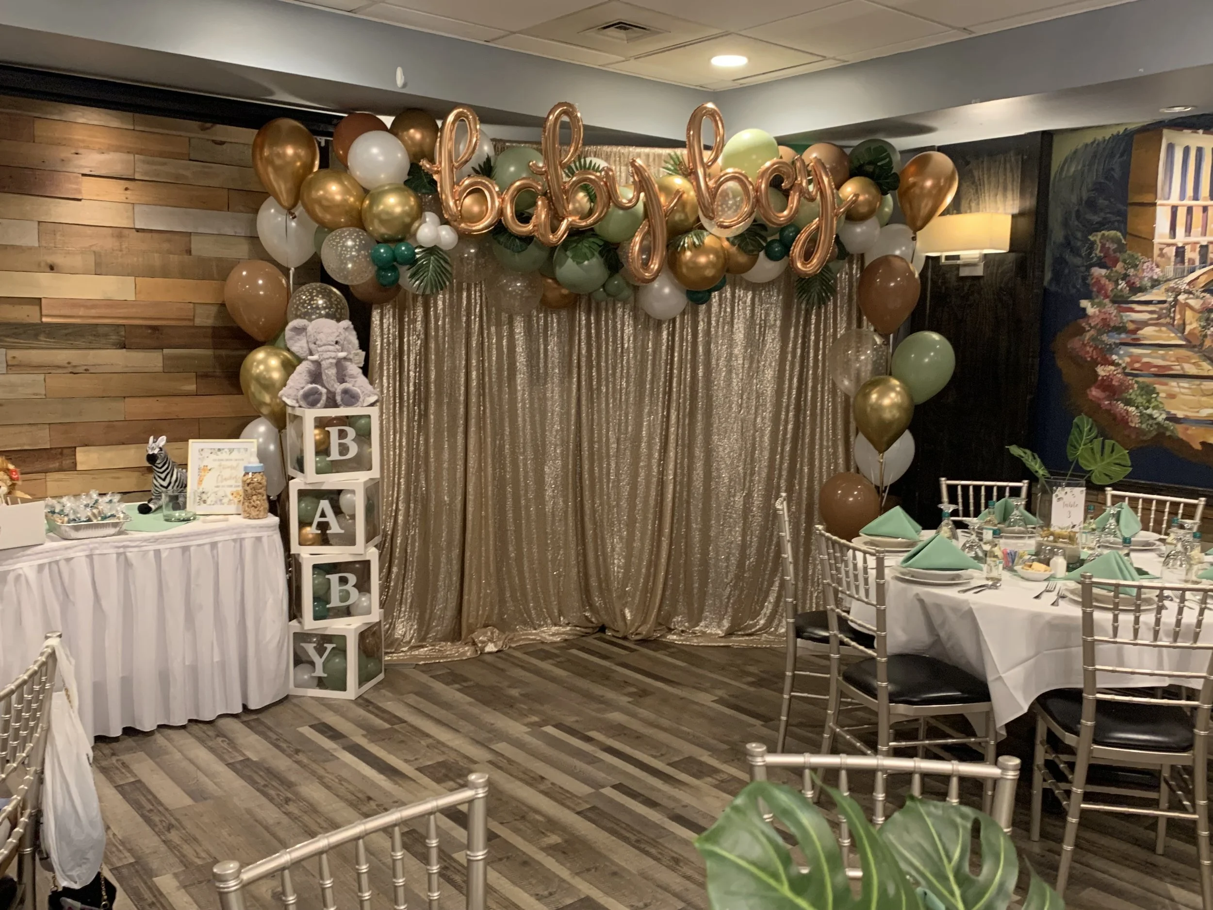 Decorations for a baby celebration with balloons, a gold curtain backdrop, and a table with party favors and stuffed animal toys.