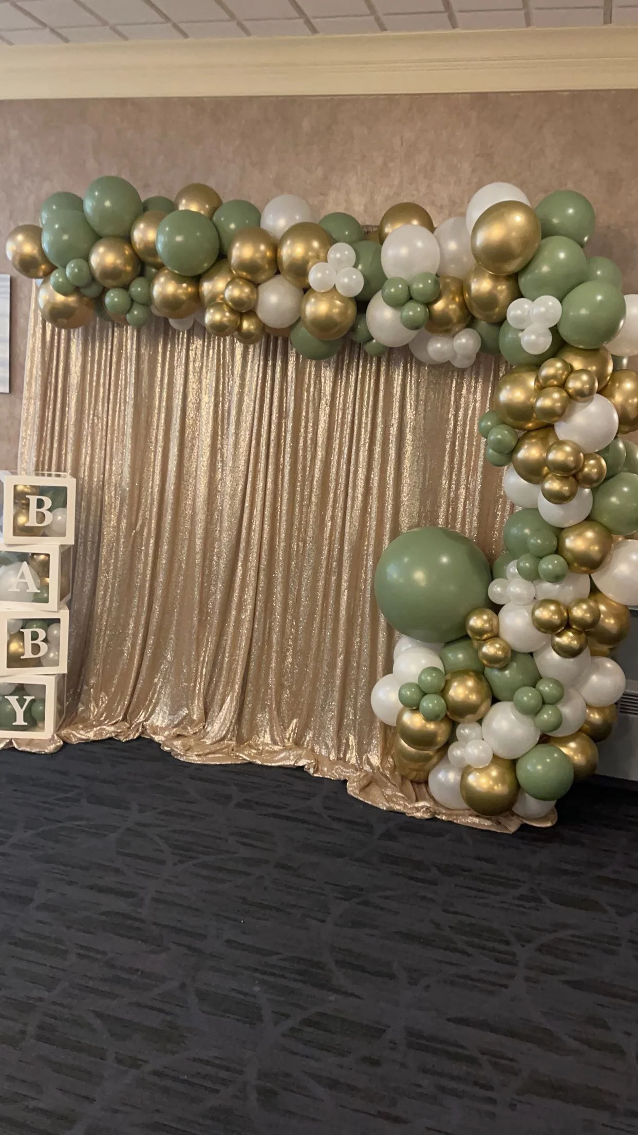Decorative balloon arch with gold, white, and green balloons in front of a gold shimmering curtain, with white blocks spelling out 'BABY' on the left.