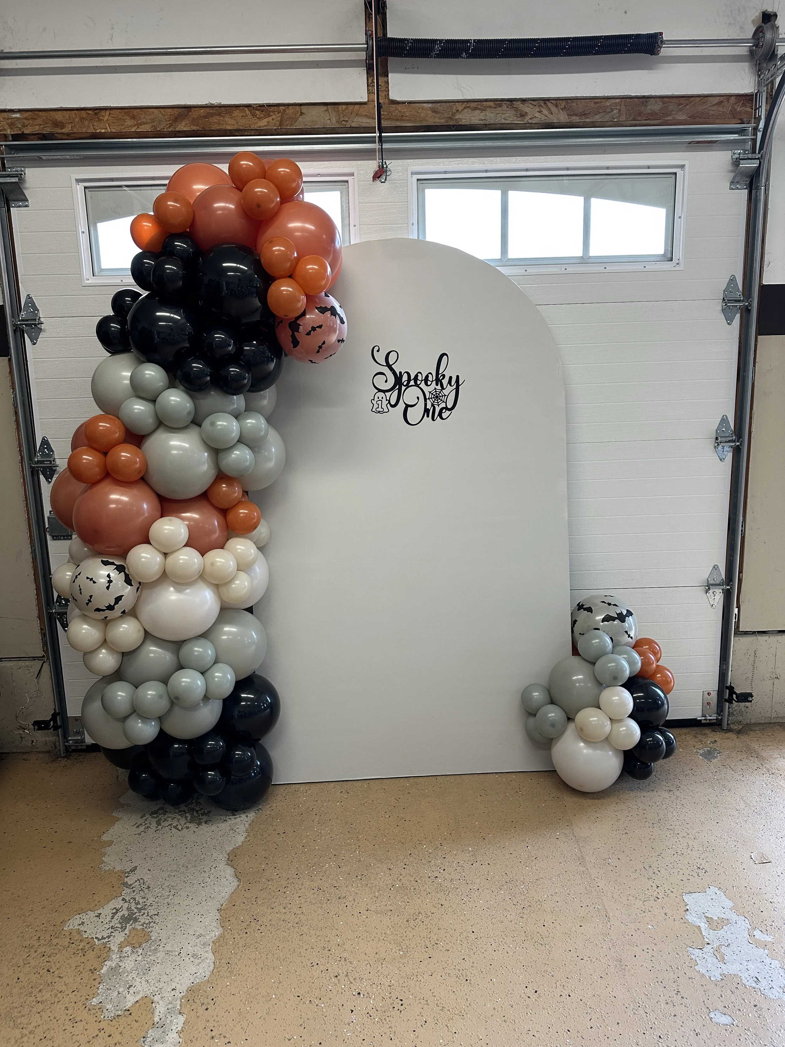 Halloween-themed balloon decorations on a white backdrop with a sign that reads 'Spooky One', featuring black, orange, white, and gray balloons, including some with bat designs.