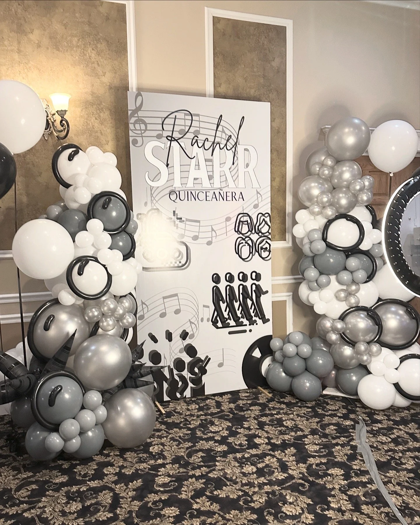Decorative balloon arrangements in black, white, silver, and gray with a large white sign that reads 'Rachel STARR QUINCEAÑERA' and musical notes, speakers, and people graphics in the background.