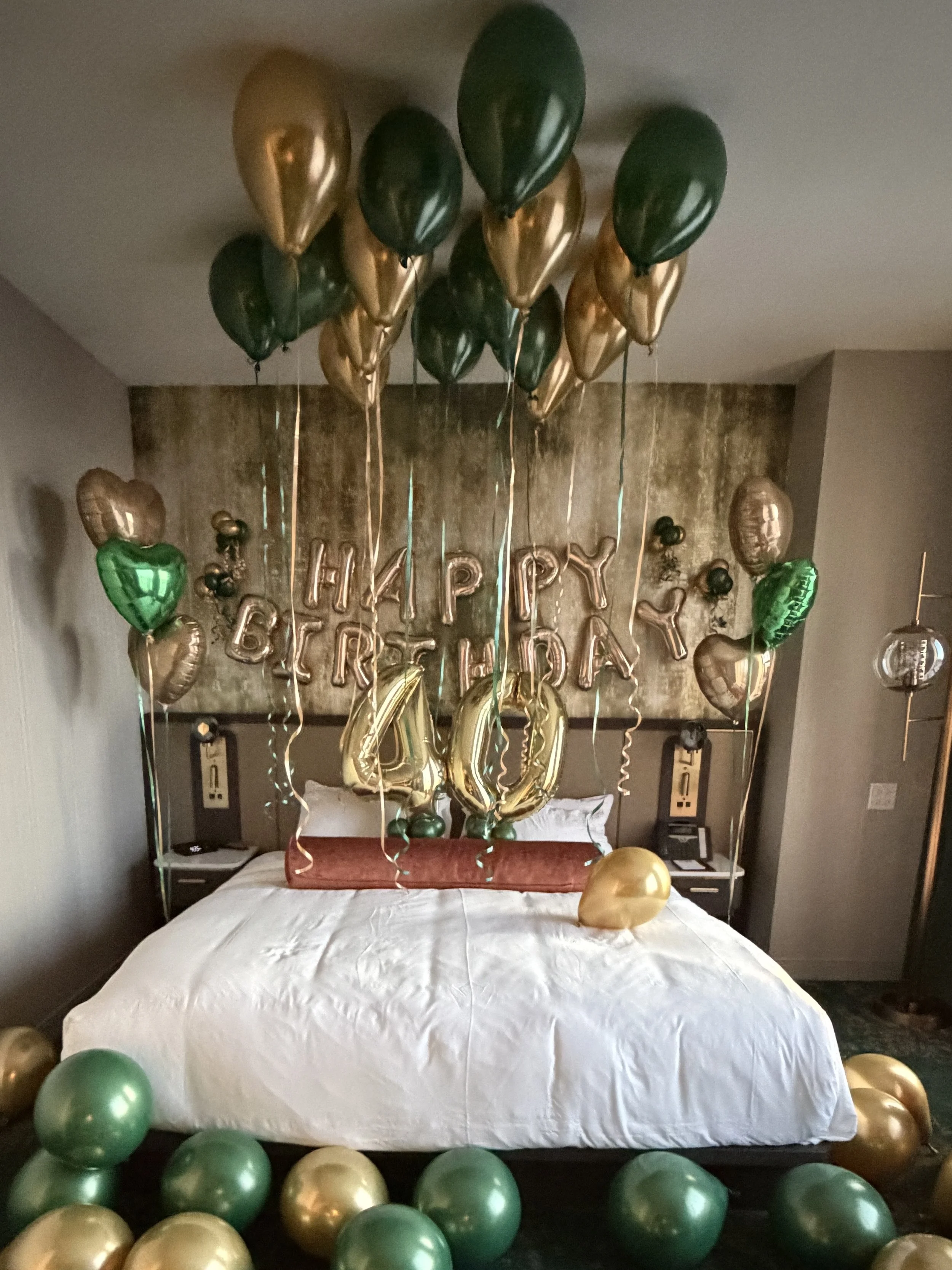 Decorated hotel room with a large bed, gold, green, and black balloons, and a 'Happy Birthday 40' sign on the wall.
