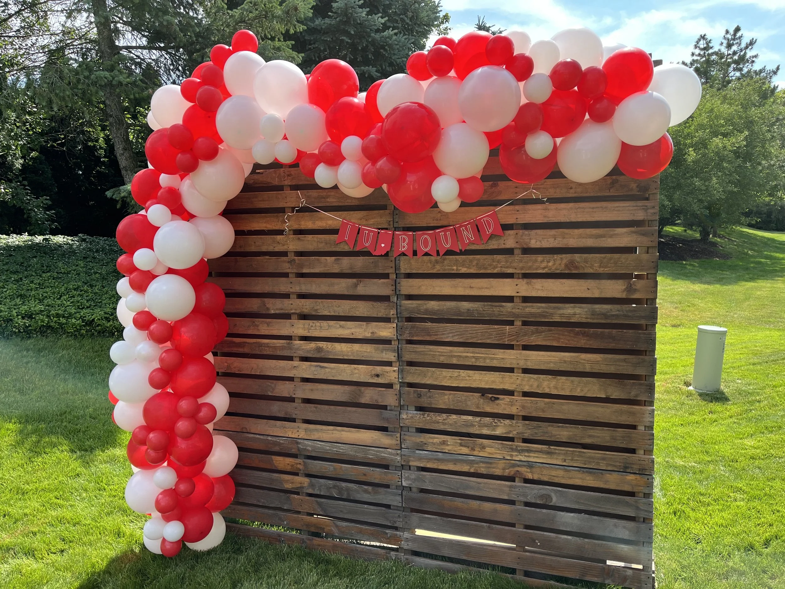 Wooden backdrop decorated with red and white balloons arranged in an arch, with a small banner reading 'HUG BOUND' hanging on the front.