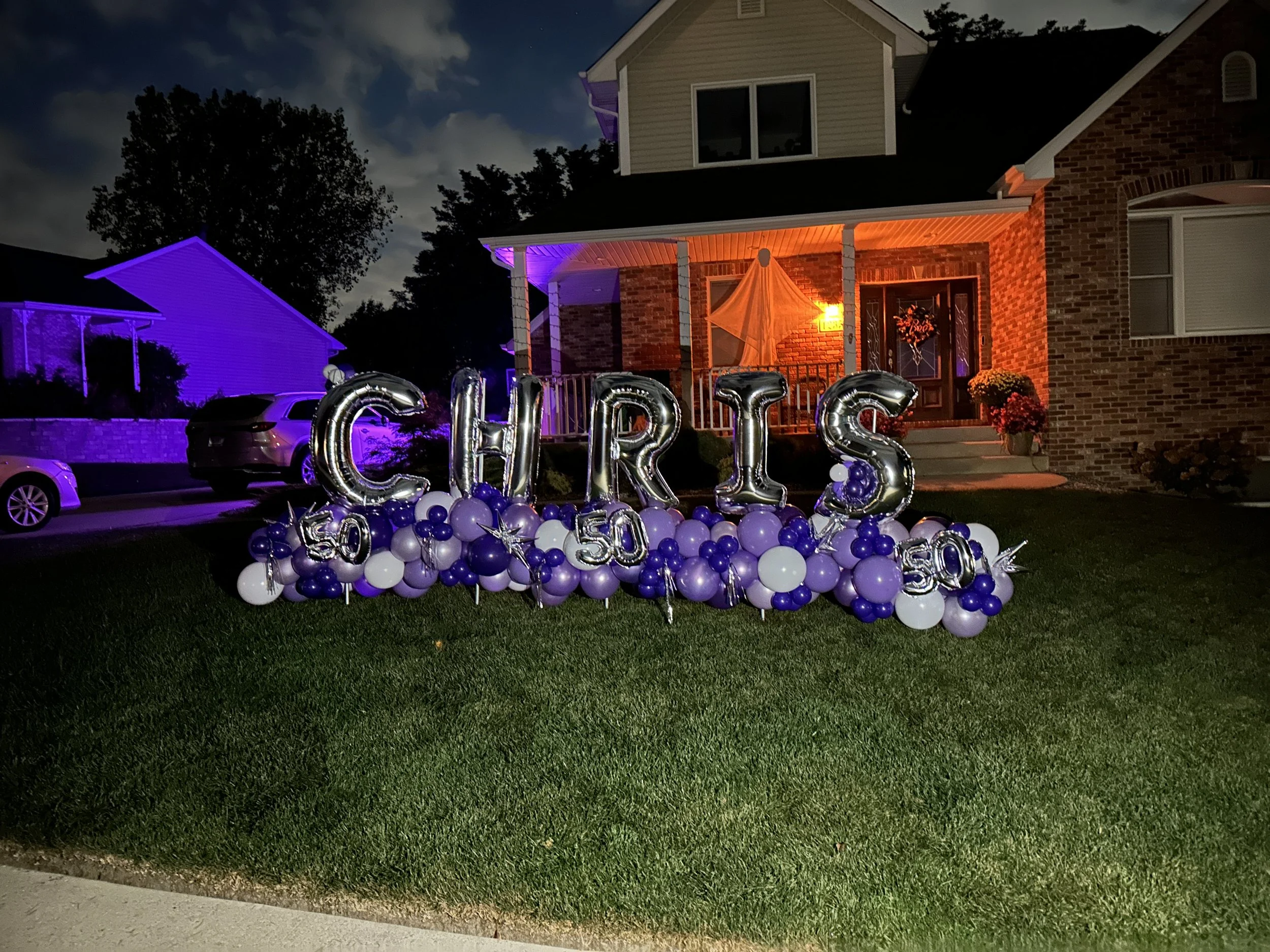 Decorative balloons spelling 'CHRIS' with 50 and 51 in the numbers, placed in front of a house decorated for Halloween at night.