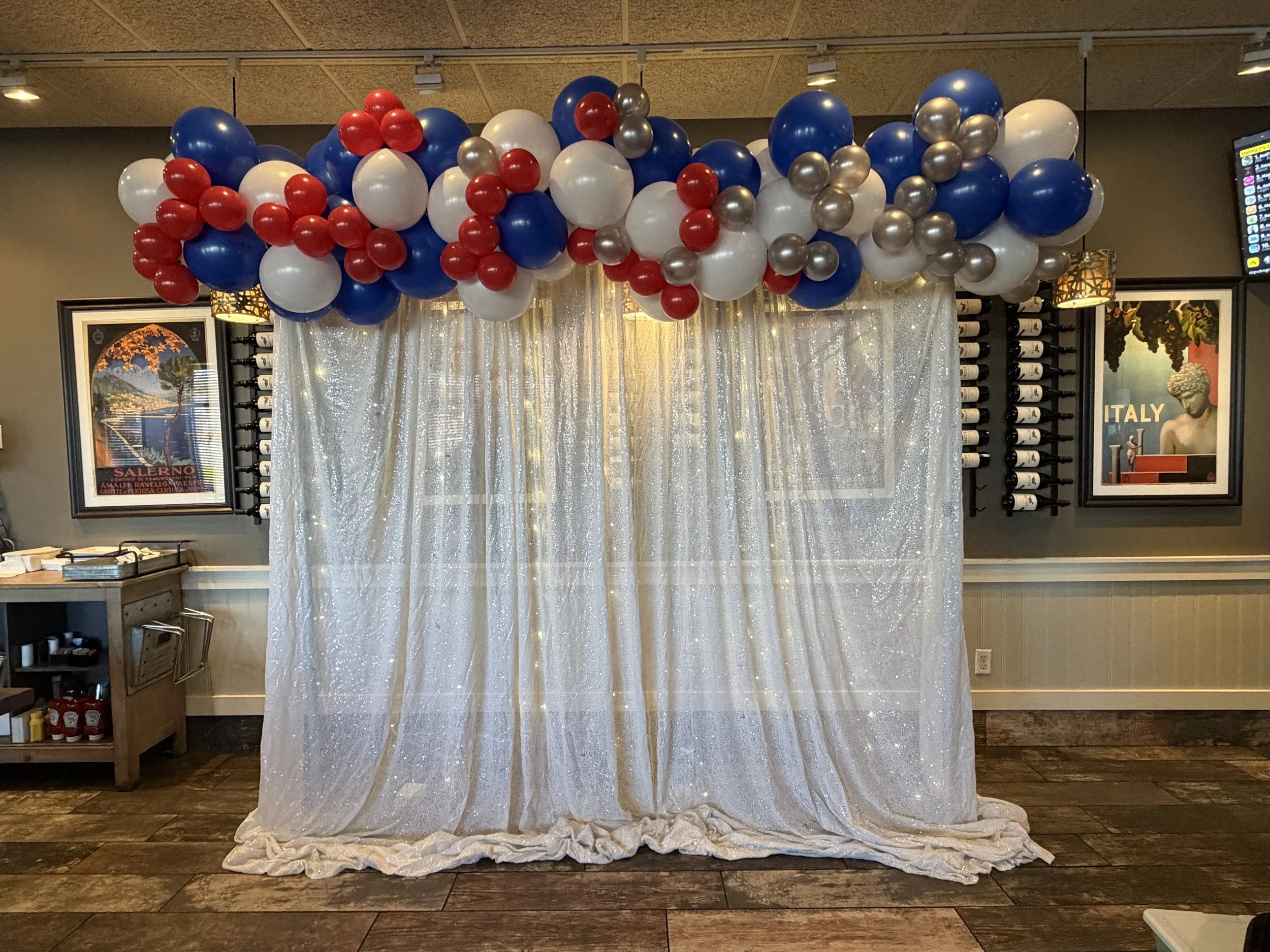 Decorative backdrop with a sheer, shimmery curtain and a balloon arch featuring red, white, blue, and silver balloons, set in a room with framed posters and a wine rack.