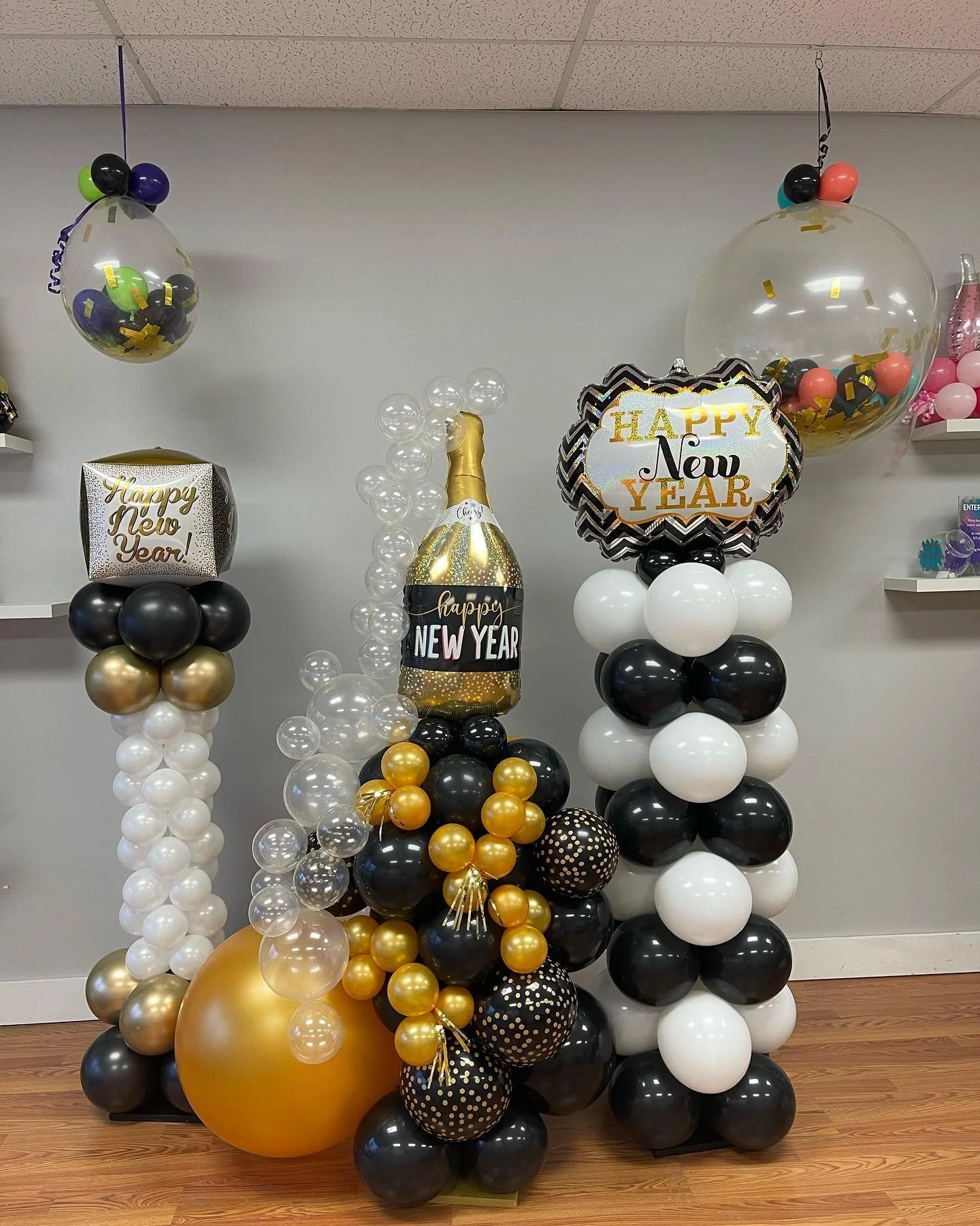 Decorative balloon arrangements with New Year and celebration themes, including a large champagne bottle-shaped balloon, smaller balloons in black, white, gold, and clear, themed balloons with 'Happy New Year' and 'HAPPY New Year' text, and clear bal