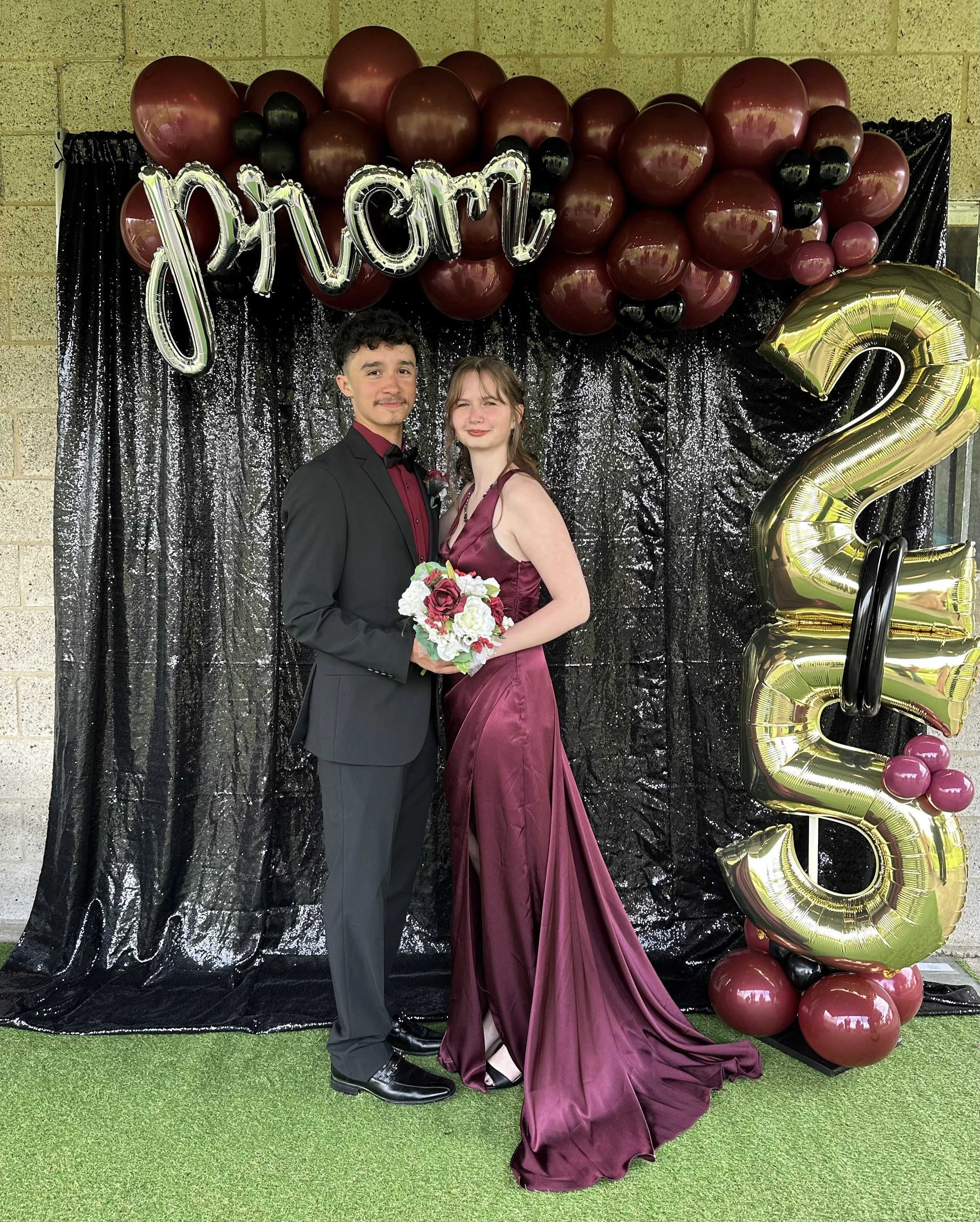A young man and woman dressed in formal attire standing together, smiling at the camera, at a prom celebration. The woman is holding a bouquet of flowers, and they are standing in front of a black sequin backdrop decorated with balloons spelling 'pro