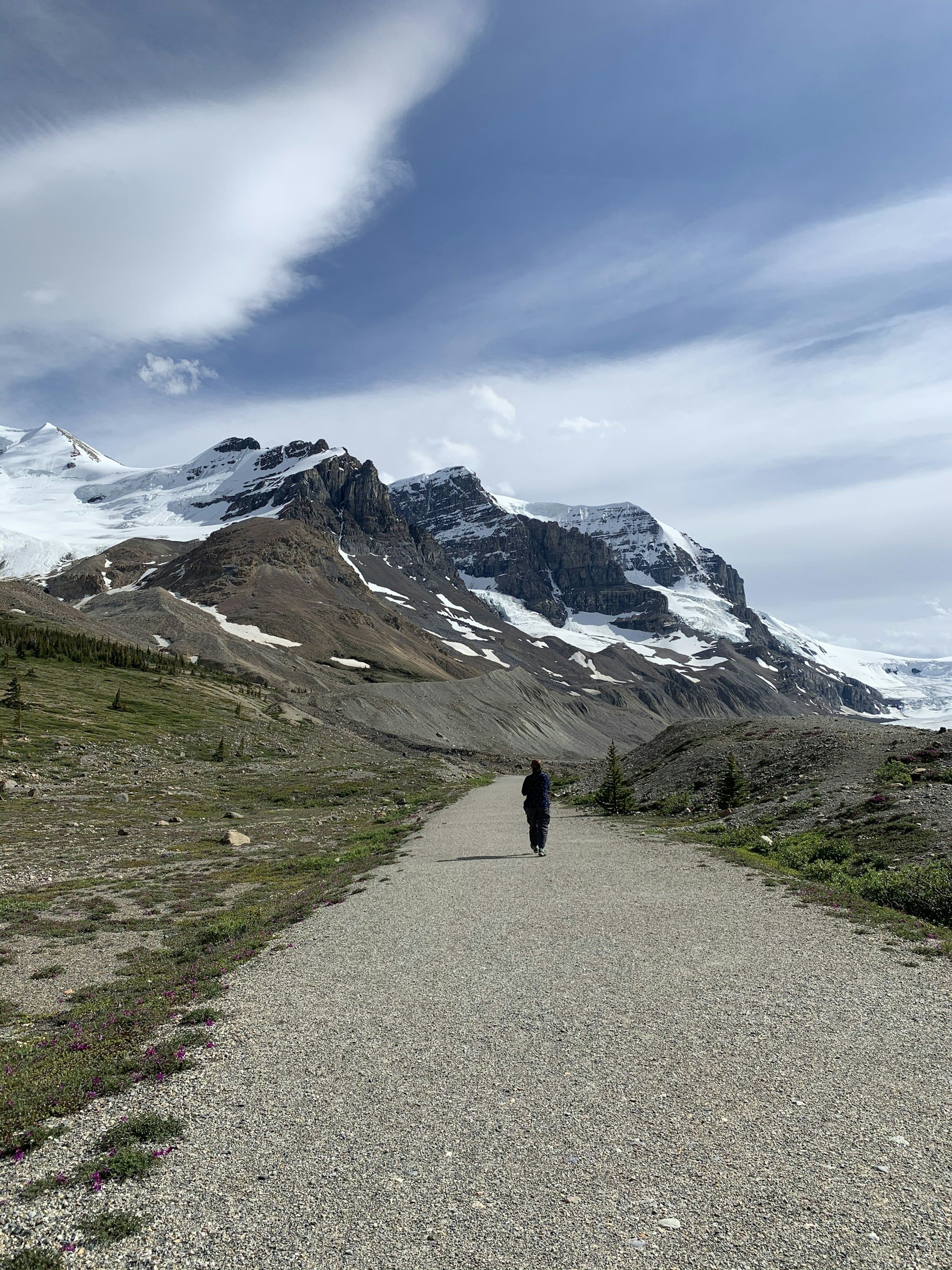 A person walking on a gravel trail through a mountainous landscape with snow-capped peaks and a cloudy sky.
