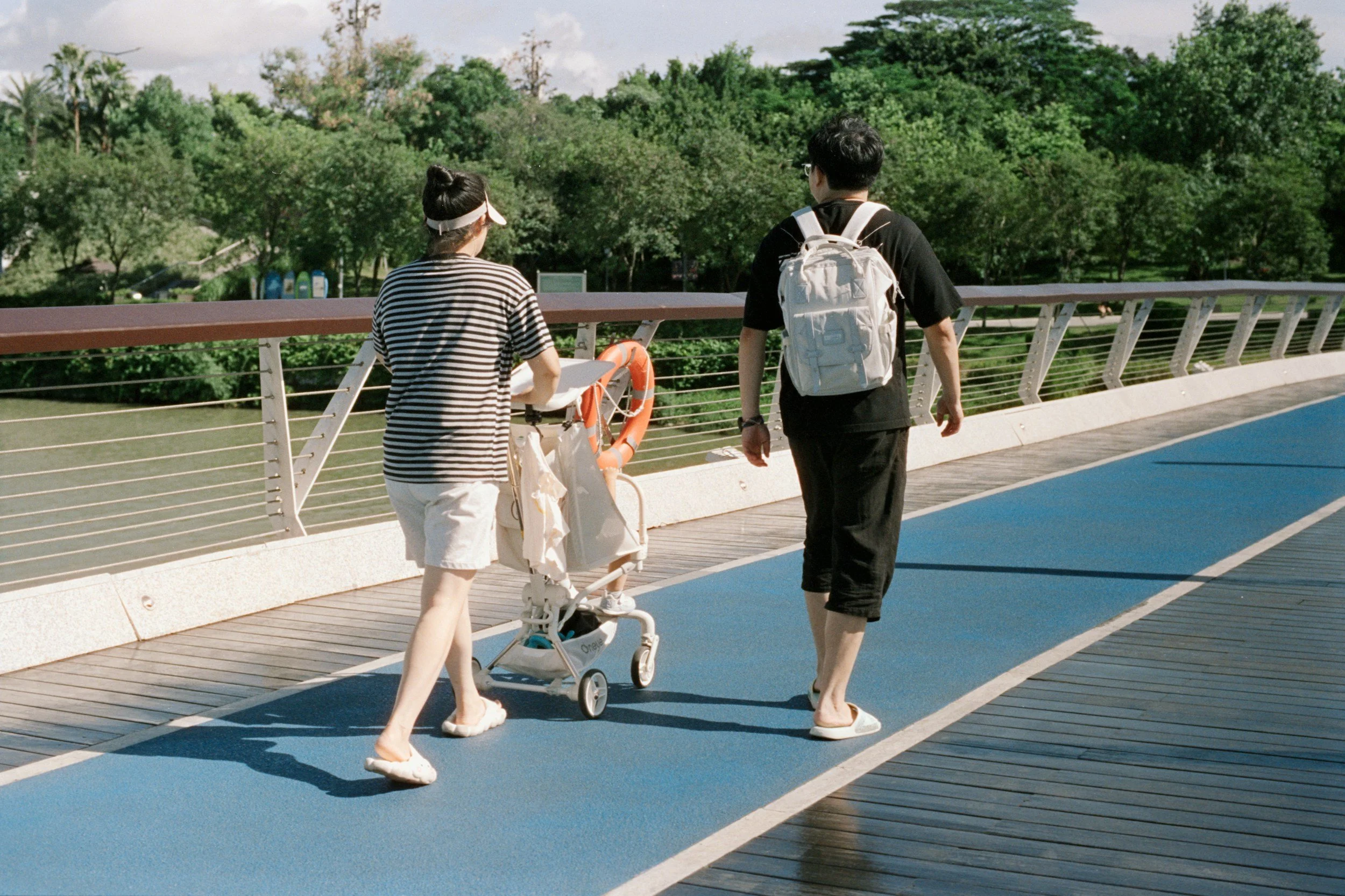 Two people walking on a bridge with lush green trees in the background. One person is pushing a stroller, and they are engaged in conversation.