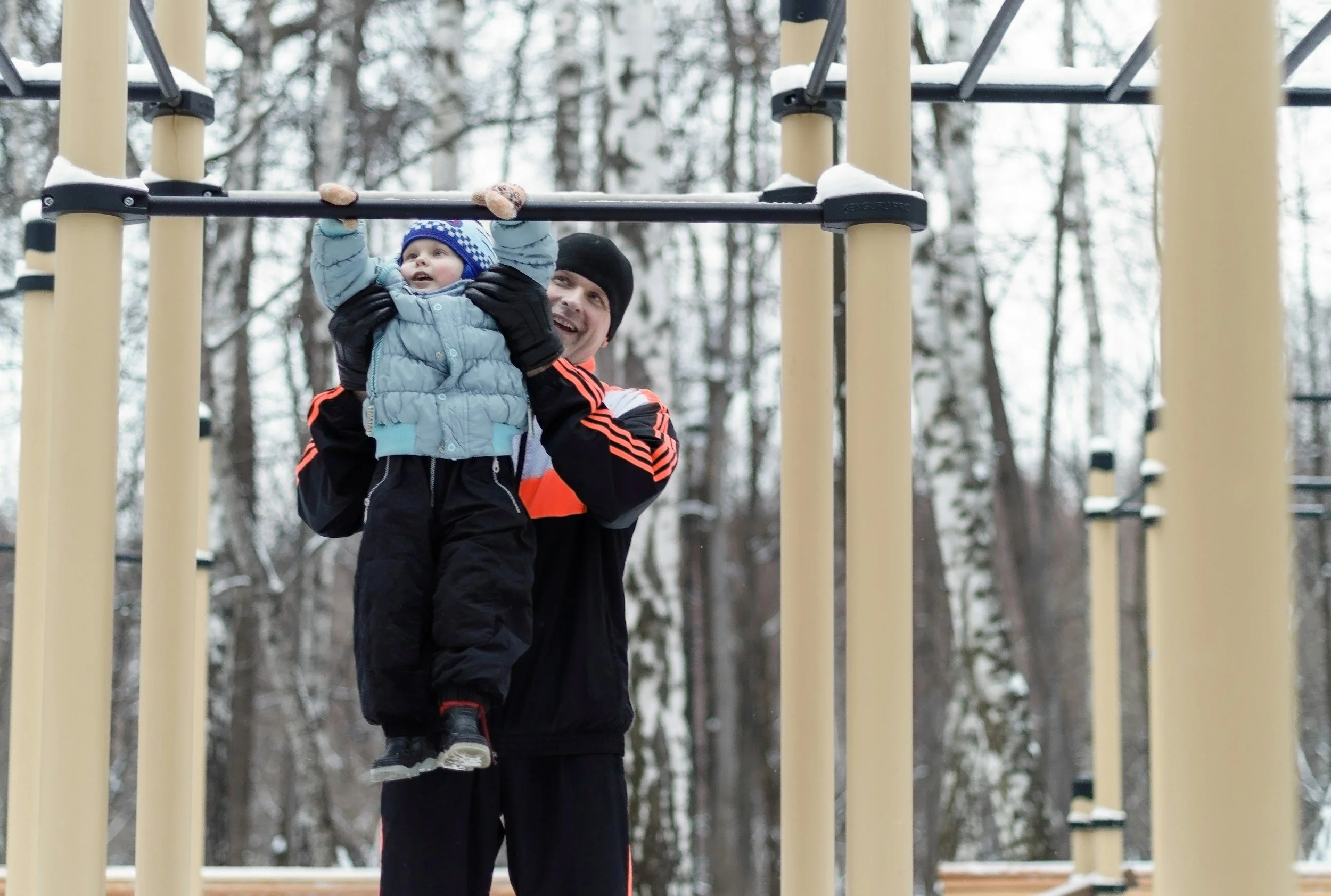 A man holding a young child on a playground structure amid snowy trees.