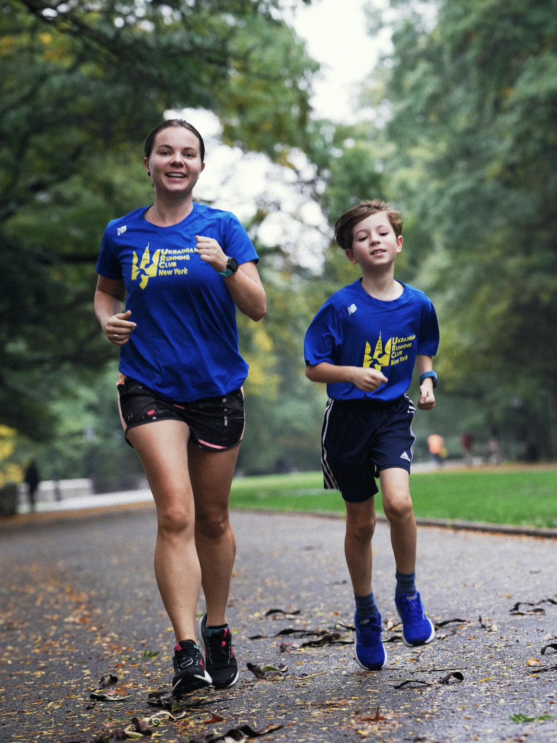 Two children, a girl and a boy, are running together on a park pathway surrounded by trees during daytime. Both are wearing blue T-shirts with yellow and white graphics and text that say 'Ukrainian Running Club New York.' The girl has short dark hair and is wearing black shorts, while the boy has light brown hair and is wearing navy blue shorts and sneakers.