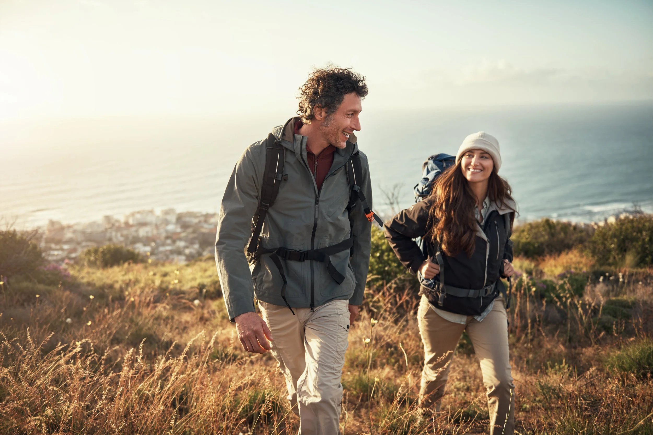 A man and a woman hiking together outdoors with a view of the ocean and city in the background, smiling and enjoying the walk.
