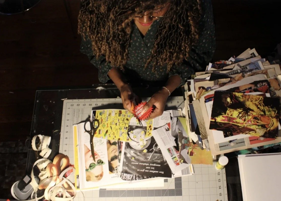 A woman with curly hair sitting at a desk covered with magazines, scissors, and tape, working on a new piece of collage art.