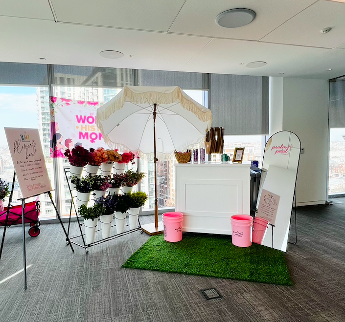 A floral shop display with white flower buckets, a white parasol with fringe, a white counter, pink trash bins, a mirror, and decorative signs on an artificial grass mat inside a modern building with large windows.