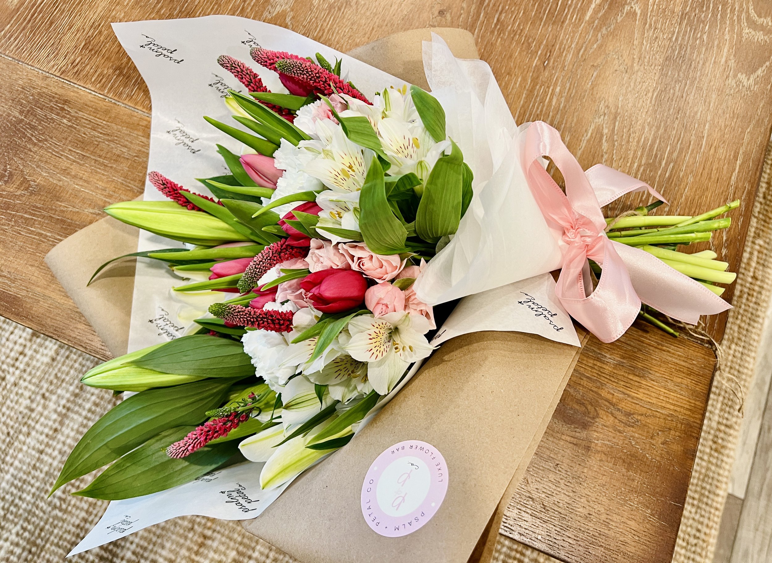 A colorful bouquet of fresh flowers including lilies, tulips, and other blooms, wrapped with tissue paper and a pink ribbon, placed on a wooden surface.